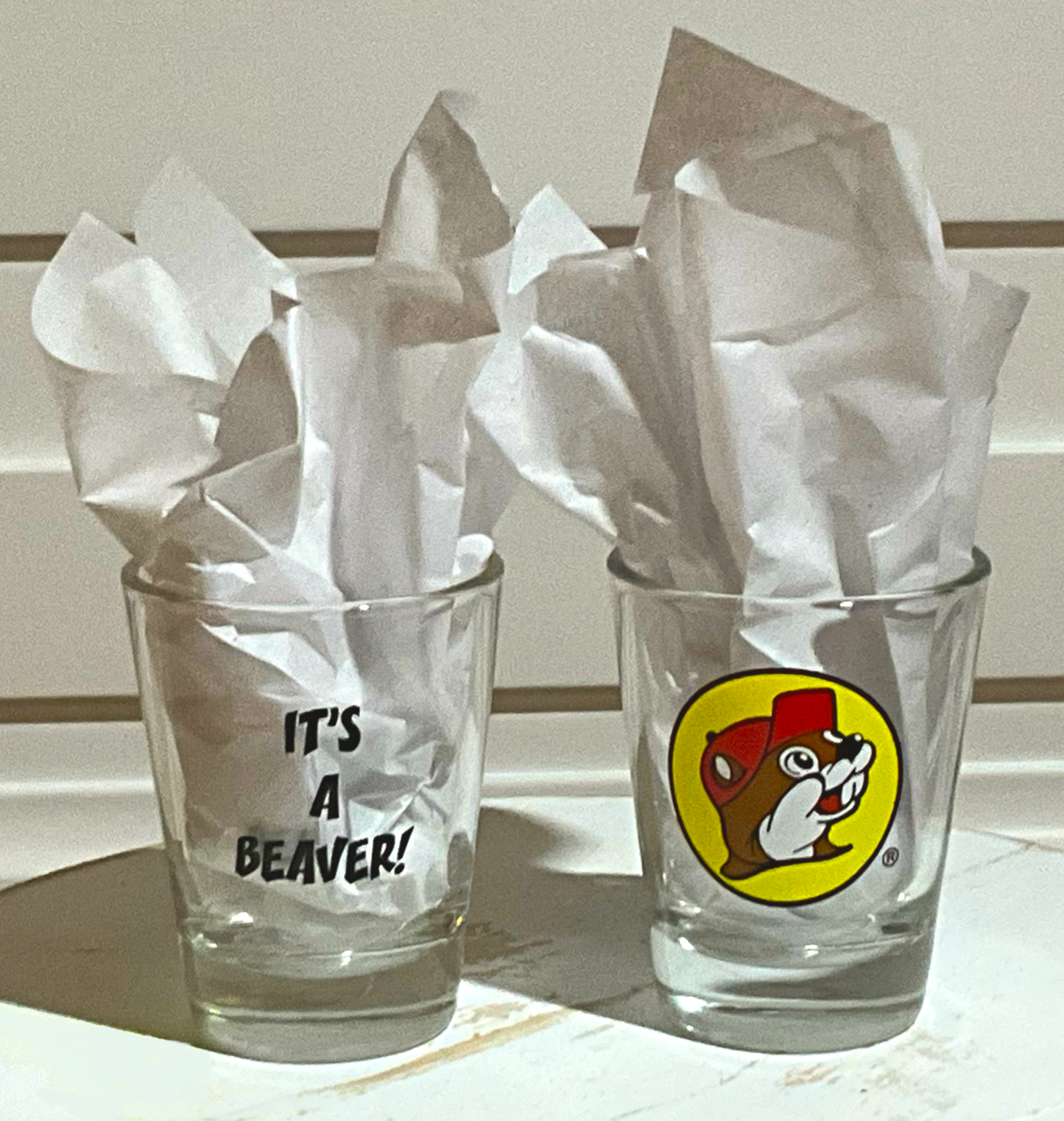 A picture of two shot glasses to demonstrate the look of the It's a Beaver Shot Glass better. On the left, the back of the glass is displayed, which has black text with IT'S A BEAVER!. On the right, is the front of the shot glass, with the Buc-ee The Beaver logo on it. Both glasses have paper in them to better illustrate the text/icon. The glasses are sitting on a white wooden bench, and a white wooden background frames them both.