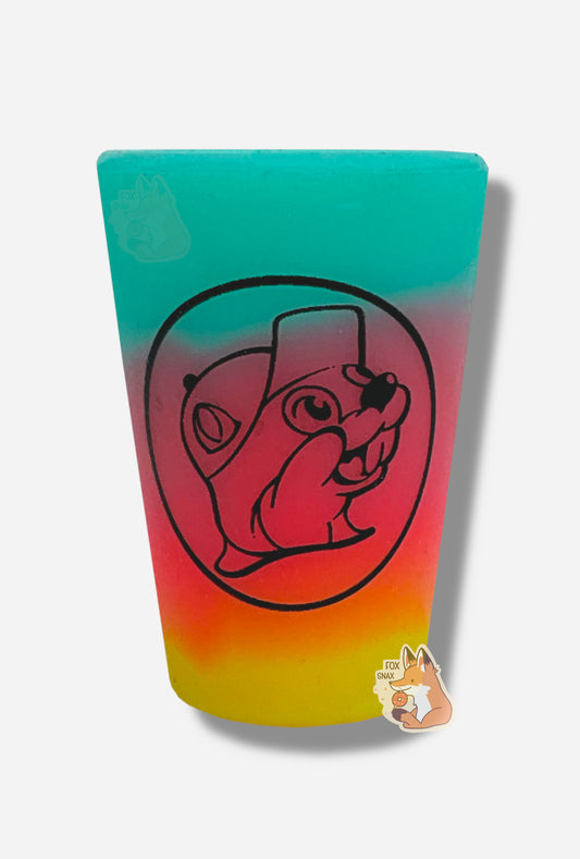 A picture of shot glass pint glass.  The glass is made of silicone, instead of actual glass, and starts with a blue/green gradient at the top, which fades to purple, then red in the middle, which fades to orange, then yellow at the bottom.  The front of the pint glass has, in a black outline, the Buc-ee's logo - a cheerful beaver wearing a ball cap.