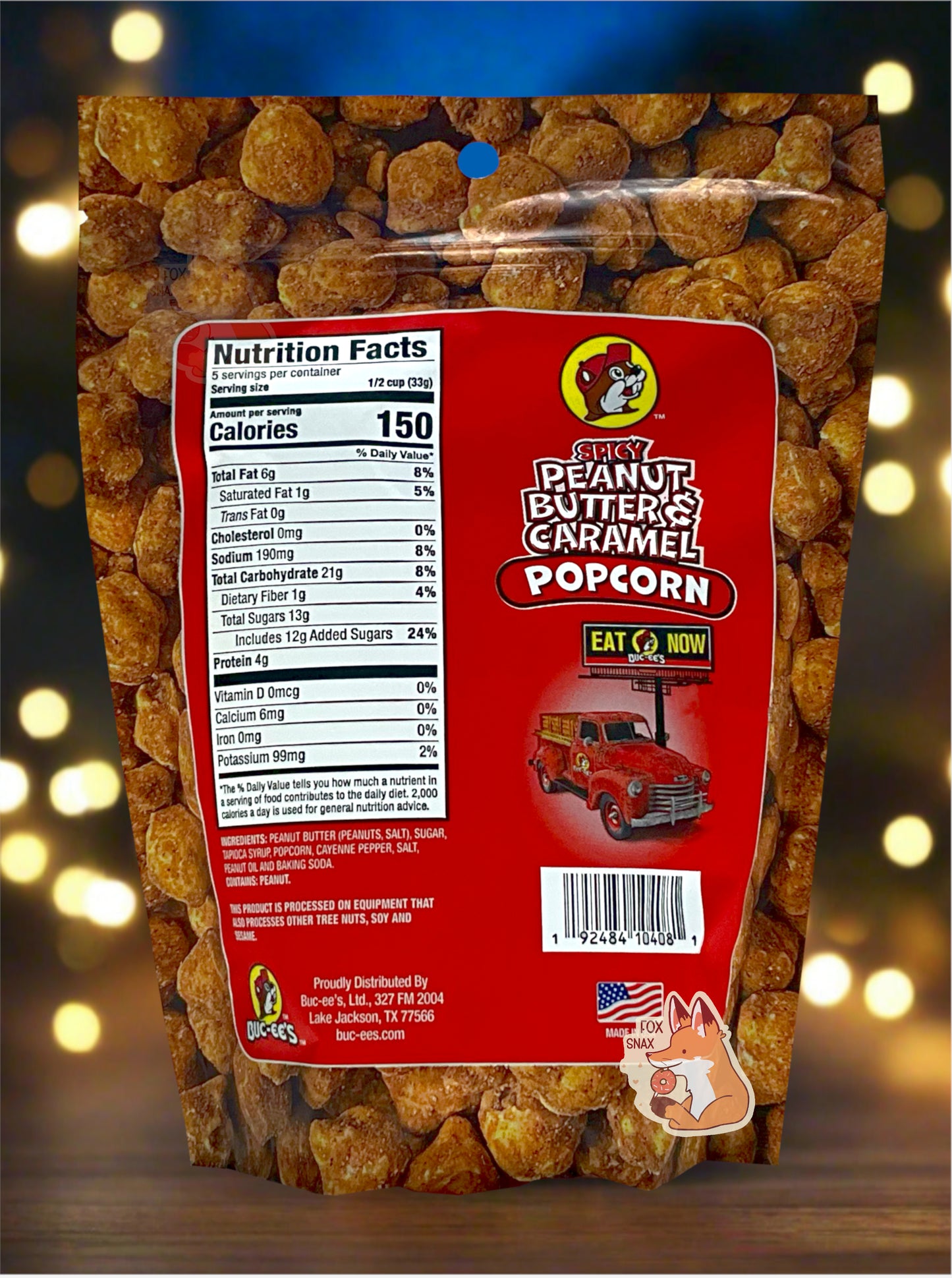 A picture of the back of a red bag of Buc-ee's Spicy Peanut Butter & Caramel Popcorn. The background is almost solely popcorn nuggets, each coated in brown peanut butter and caramel coating. The right side reads SPICY PEANUT BUTTER & CARAMEL POPCORN and has a picture of a red truck under a Buc-ee's EAT NOW sign. Underneath that is the American flag, and says MADE IN USA. The bag is sitting on a table, outdoors at night; a dark blue sky can be seen against string lights.