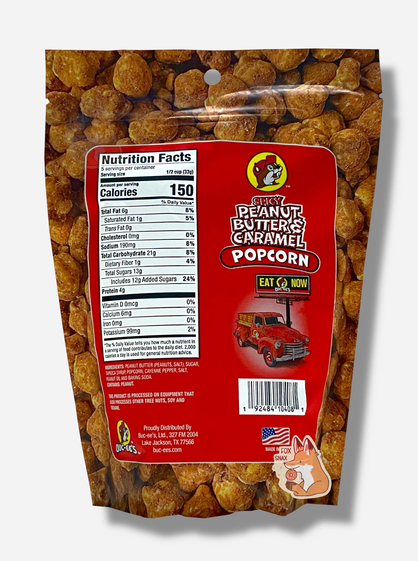 A picture of the back of a red bag of Buc-ee's Spicy Peanut Butter & Caramel Popcorn. The background is almost solely popcorn nuggets, each coated in brown peanut butter and caramel coating. The right side reads SPICY PEANUT BUTTER & CARAMEL POPCORN and has a picture of a red truck under a Buc-ee's EAT NOW sign. Underneath that is the American flag, and says MADE IN USA.