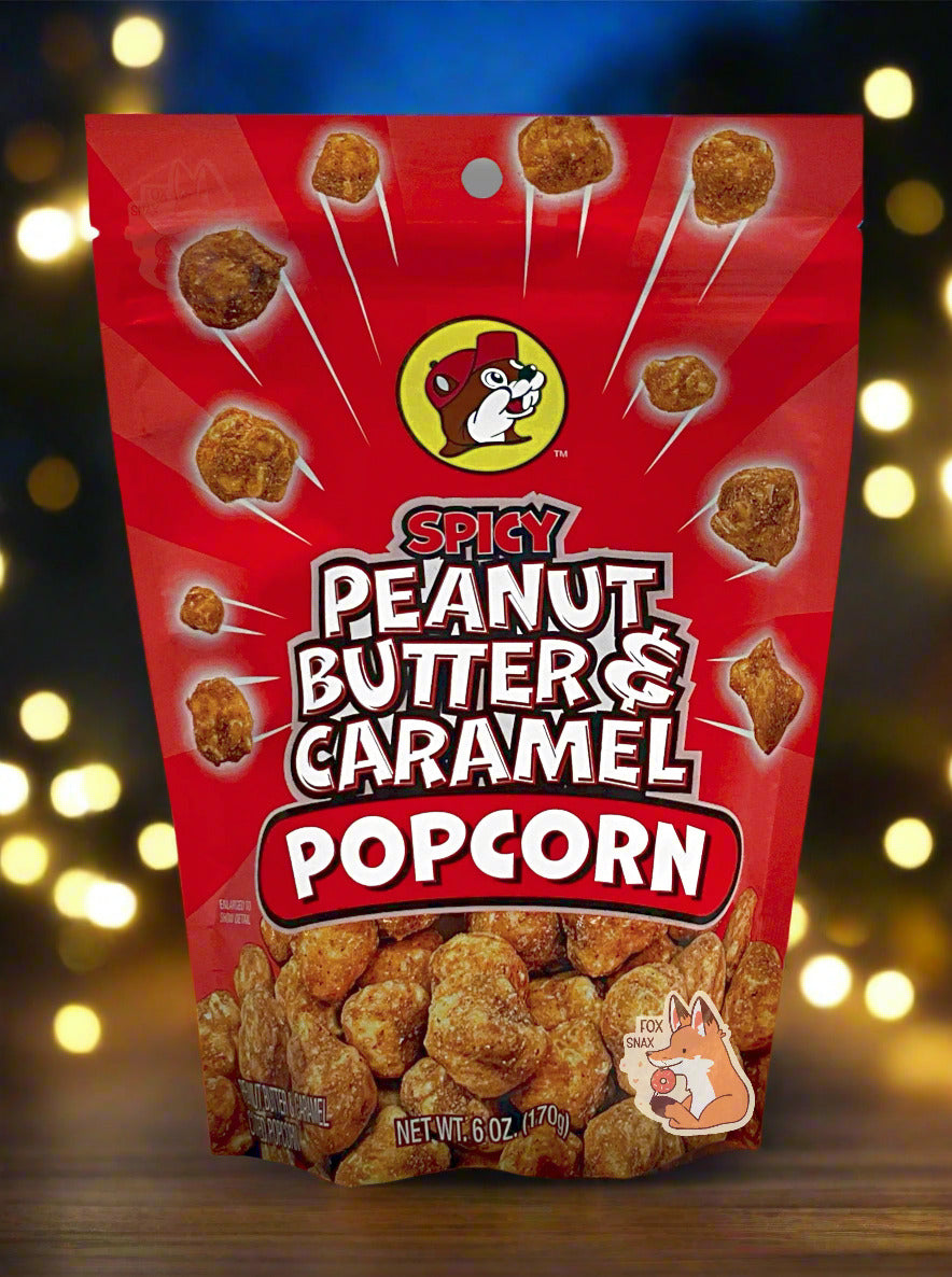 A picture of a red bag of Buc-ee's Spicy Peanut Butter & Caramel Popcorn. The front of the bag has a group of popcorn nuggets, each coated in brown peanut butter and caramel coating, exploding from the center. A pile of popcorn is on the bottom. The top center has the Buc-ee's logo, with cheerful beaver wearing a red ball cap, framed in a yellow circle, and reads SPICY PEANUT BUTTER & CARAMEL POPCORN. The bag is sitting on a table, outdoors at night; a dark blue sky can be seen against string lights.