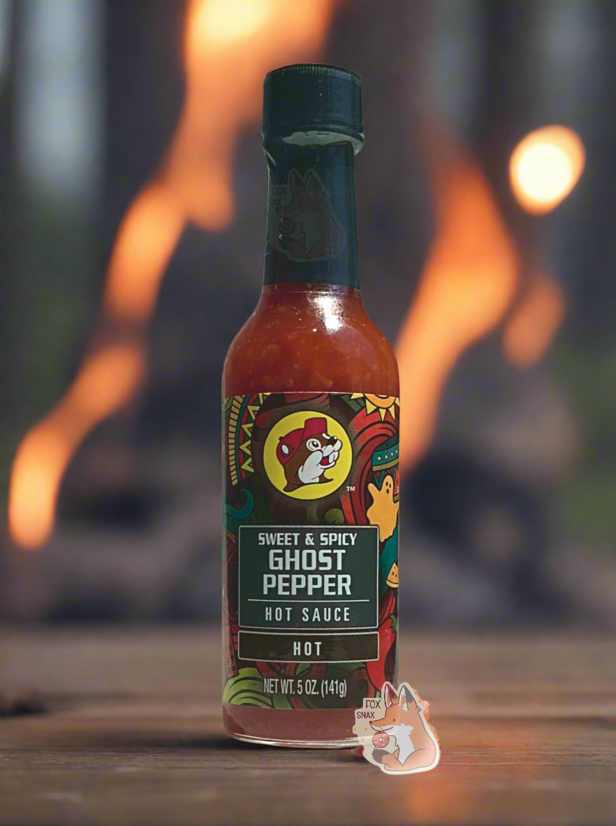 A picture of a small, tall glass bottle of hot sauce. The top of the bottle is sealed with a plastic top. The front label of the bottle reads SWEET & SPICY GHOST PEPPER HOT SAUCE, HOT. The Buc-ee's logo can be seen on the front; the background is a fairly busy abstract artwork featuring an orange ghost, sun, and several colorful abstract shapes. The sauce inside is dark red. The bottle is sitting on a flat piece of wood; a large campground fire can be seen in the background.
