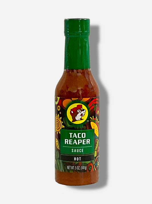 A picture of a small, tall glass bottle of hot sauce.  The top of the bottle is sealed with a plastic top.  The front label of the bottle reads TACO REAPER HOT SAUCE, HOT.  The Buc-ee's logo can be seen on the front; the background is a fairly busy abstract artwork featuring a cactus, two maracas, a lemon, a taco, and a red pepper.  The sauce inside is dark orange.