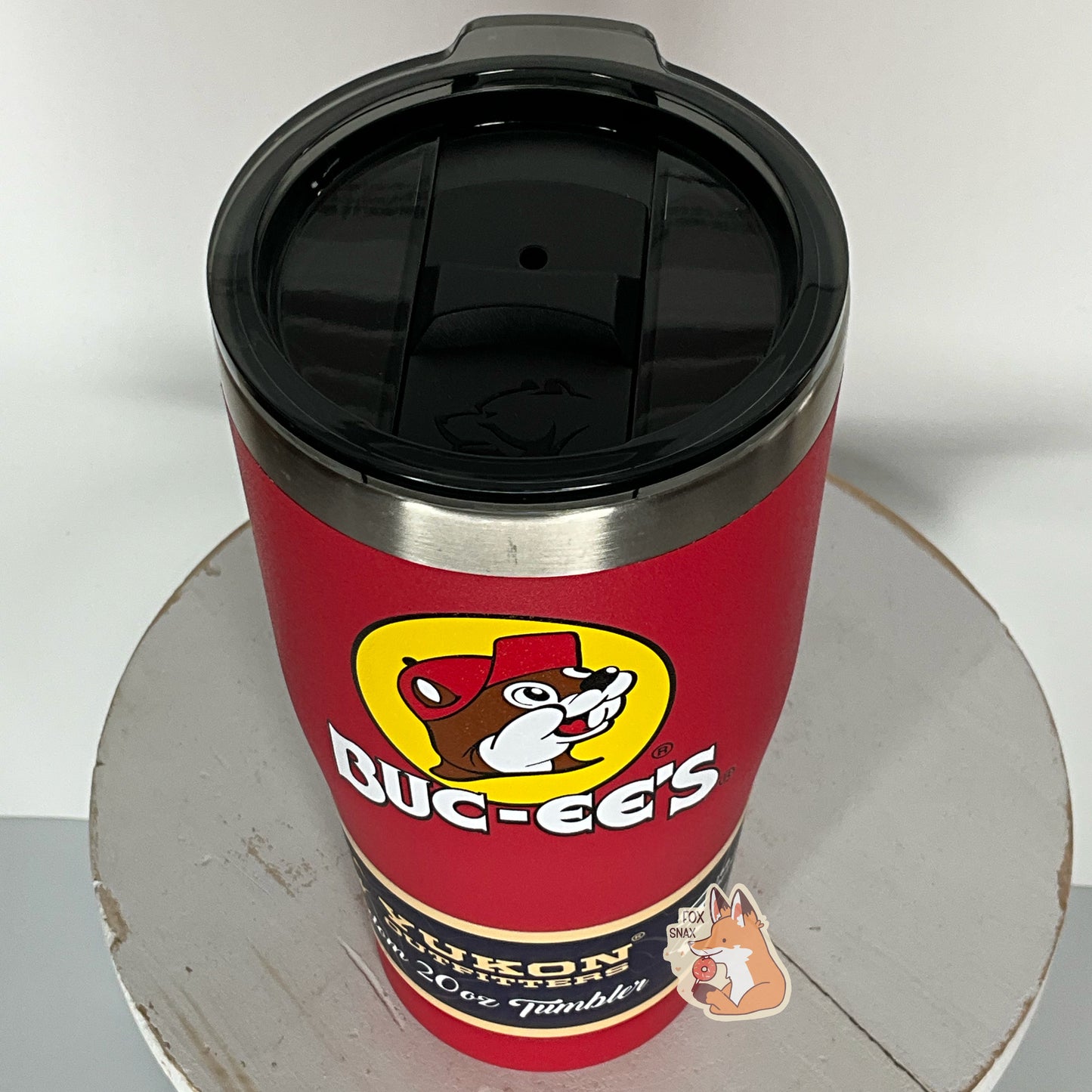 A three-quarters view of a picture of a tall, red, tumbler, with a big picture of Buc-ee The Beaver on it. The surface is smooth and red, as it is red paint covering stainless steel. There is no handle. The top is black plastic, and shows a slider in the middle with the imprint of the Yukon bear's head on it. The top of the mug is lined with a stainless steel band. A paper wrapper around the bottom reads: Yukon Outfitters. Freedom 20 oz Tumbler. The tumbler is sitting on a white circular stool.