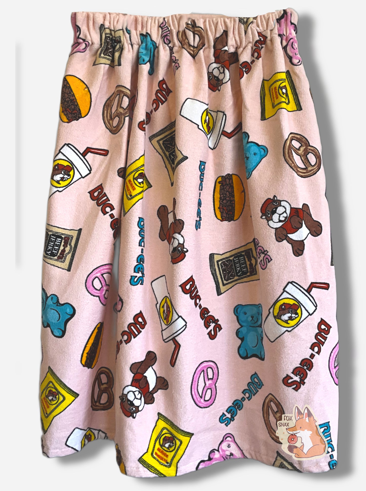 A picture of the back of a pink towel wrap. The wrap looks fluffy, and has various rotated Buc-ee The Beaver on it, beaver nuggets, a pretzel, the words BUC-EES, a brisket sandwich, a white drink container, and a pink gummy bear icons all over it.