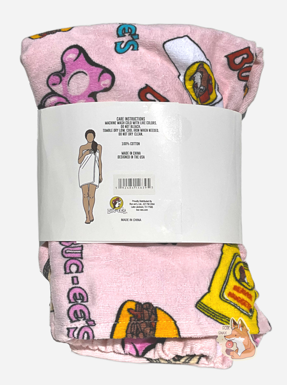 A picture of the back of a wrapped pink towel wrap. The wrap looks fluffy, and has a picture of Buc-ee The Beaver on it, beaver nuggets, a pretzel, and the words BUC-EES. A white paper wrap surrounds the middle of the folded wrap, and reads CARE INSTRUCTIONS, MACHINE WASH COLD WITH LIKE COLORS. DO NOT BLEACH. TUMBLE DRY LOW. COOL IRON WHEN NEEDED. DO NOT DRY CLEAN.