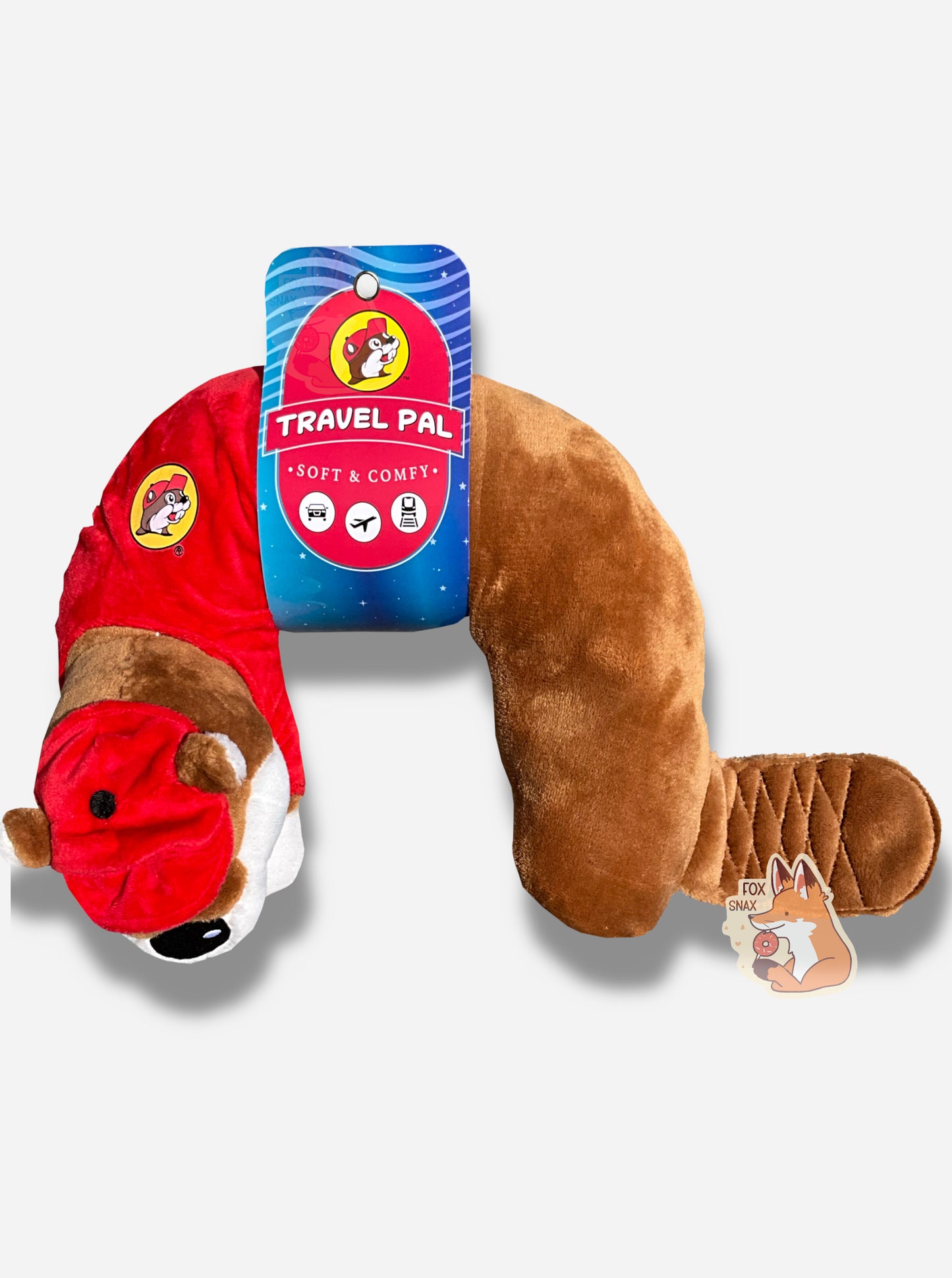 A picture of a mostly u-shaped, travel pal pillow. It is is shaped roughly like Buc-ee the Beaver. Buc-ee is wearing a red ball cap, a red Buc-ee's shirt complete with Buc-ee logo, and sports a brown beaver tail. The paper label around his midsection reads TRAVEL PAL, then SOFT AND COMFY, then shows three icons - a car, a plane, and a train/subway car.