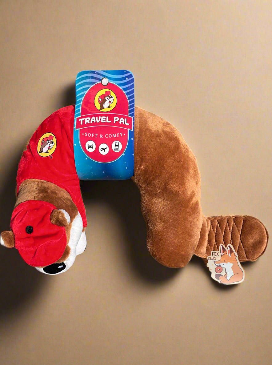 A picture of a mostly u-shaped, travel pal pillow. It is is shaped roughly like Buc-ee the Beaver. Buc-ee is wearing a red ball cap, a red Buc-ee's shirt complete with Buc-ee logo, and sports a brown beaver tail. The paper label around his midsection reads TRAVEL PAL, then SOFT AND COMFY, then shows three icons - a car, a plane, and a train/subway car. The pillow is sitting on a tan soft couch in a living room.