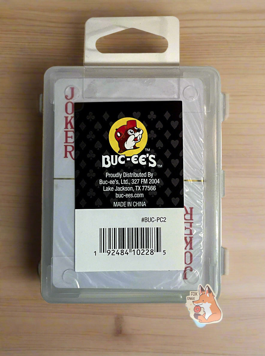 A picture of the back of a pack of Buc-ee's Waterproof Playing Cards. The pack is plastic, rectangular, and clear. A joker card can be seen through the plastic. A small rectangular sticker with the Buc-ee's logo against a hearts/spades/diamonds/clubs background can be seen. The cards are sitting on a light wood table.