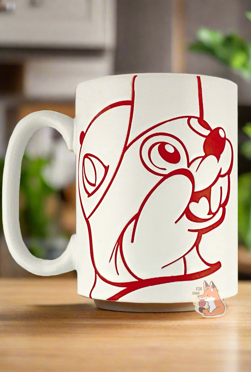 A picture of a ceramic, red and white coffee mug. The mug prominently features a picture of Buc-ee The Beaver on the front and back, in red. The interior of the mug is a bright red as well, and rest of the mug is white. The mug is resting on a pretty light wood table, against a platn-decorated kitchen background.