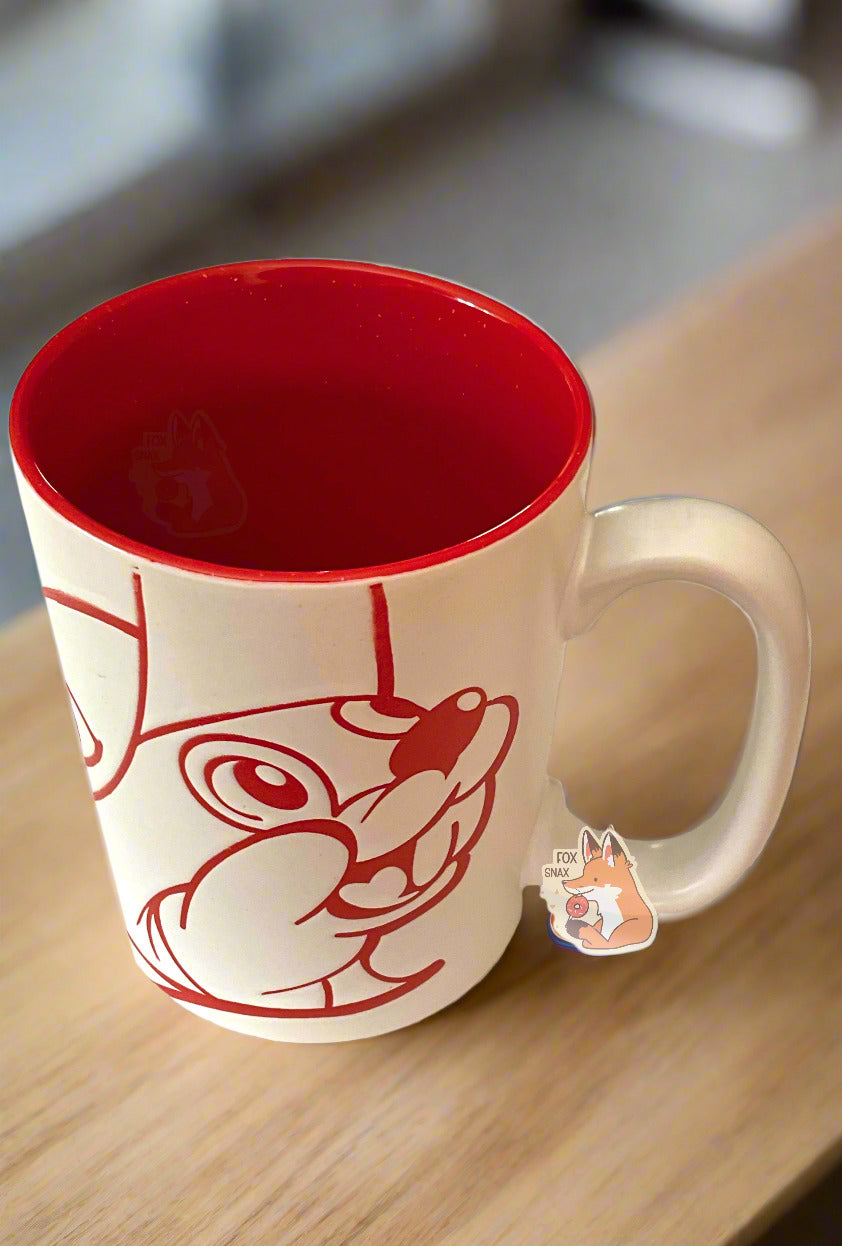 A picture of a ceramic, red and white coffee mug. The mug prominently features a picture of Buc-ee The Beaver on the front and back, in red. The interior of the mug is a bright red as well, and rest of the mug is white. The background shows the mug on a wooden rail against a warm, clean kitchen background.