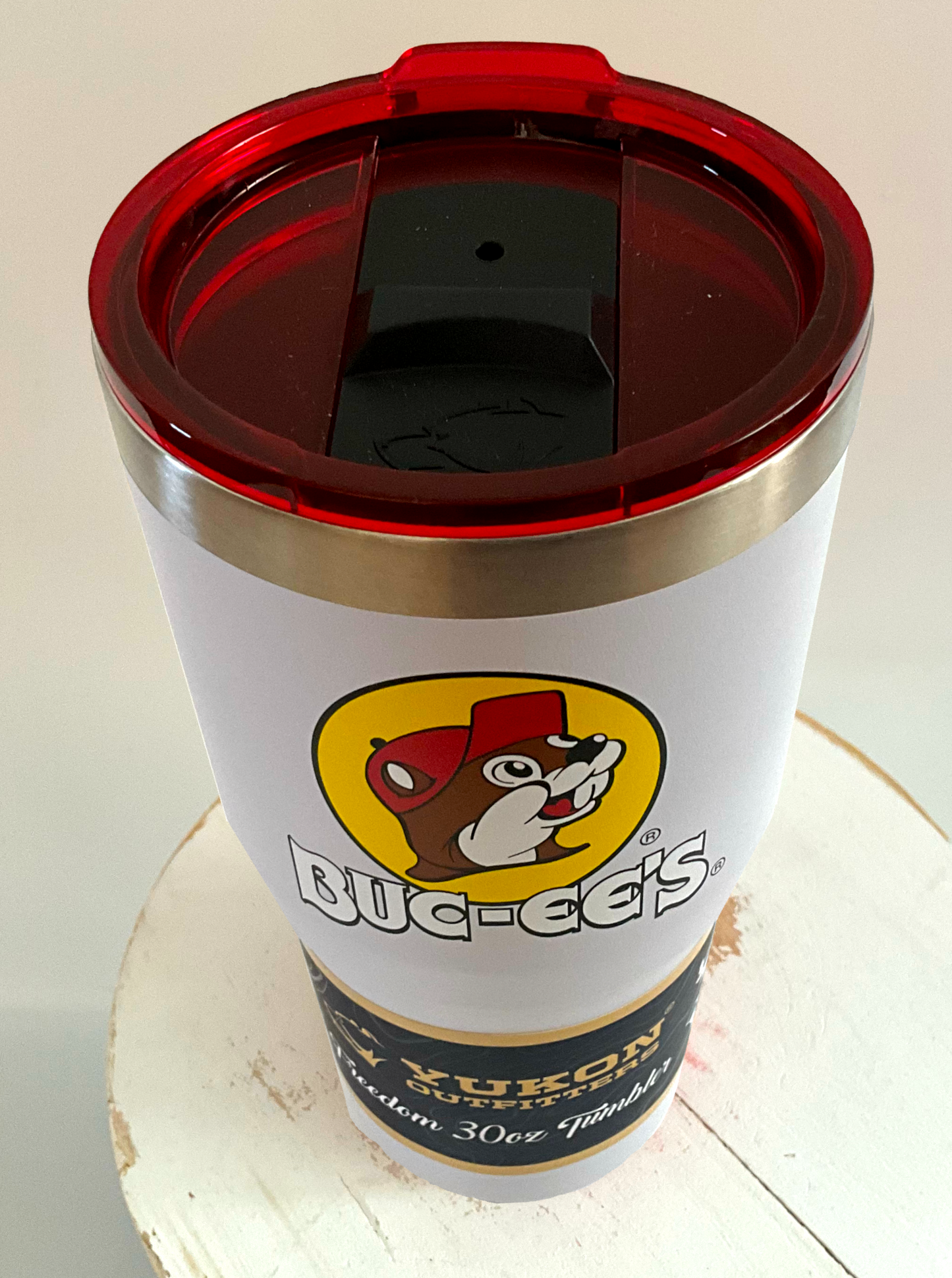 A 3/4 view of a picture of a tall, white, tumbler, with a big picture of Buc-ee The Beaver on it. The surface is smooth and white, as it is white paint covering stainless steel. There is no handle. The top is red plastic, and shows a black slider in the middle with the imprint of the Yukon bear's head on it. The top of the mug is lined with a stainless steel band. A paper wrapper around the bottom reads: Yukon Outfitters. Freedom 30 oz Tumbler. The tumbler is sitting on a white circular stool.