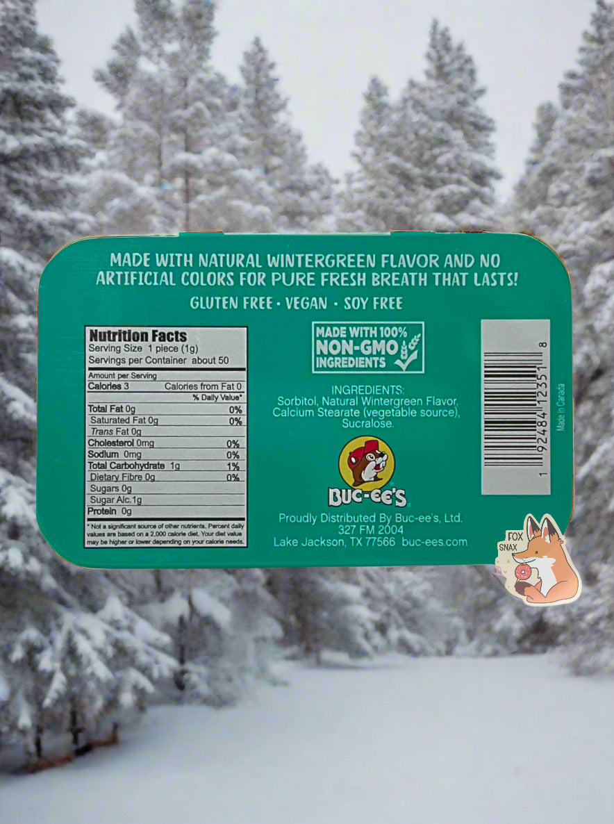 A picture of a back of a tin of Buc-ees Wintergreen Mints. The back cover is decorated with a small picture of Buc-ee The Beaver, and has the following text: Made With Natural Wintergreen Flavor And No Artificial Colors Or Flavors For Pure Fresh Breath That Lasts!, Gluten Free, Vegan, Soy Free, made with 100% Non-GMO Ingredients. The background is a series of snow-covered pine trees; a winter wonderland.