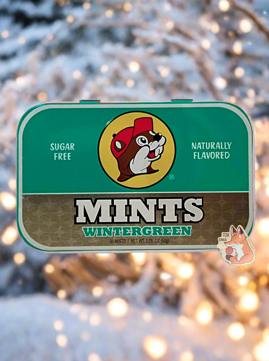 A picture of a tin of Buc-ees Wintergreen Mints. The cover is decorated with a big picture of Buc-ee The Beaver, and has a green and gold background. The tin reads Sugar Free, Naturally Flavored, Mints, Wintergreen. The background is a winter wonderland; close to the branches of a snow-laden tree, various yellowish lights can be seen.