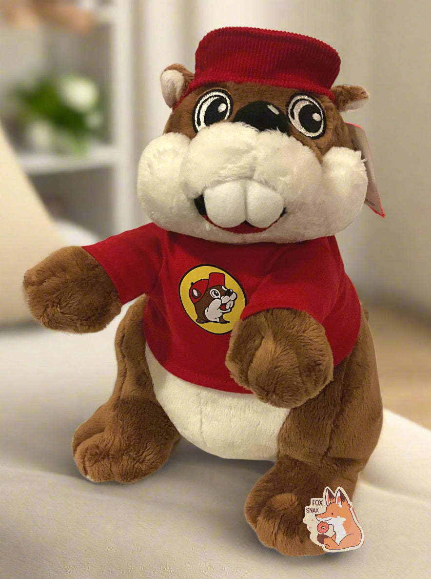 A picture of an adorable Buc-ee the Beaver plush.  The plush is of Buc-ee, wearing a red Buc-ee's shirt and red ball cap, and is looking to the left.  It is roughly a foot tall and covered in plush brown and white stuffed animal "fur".  The background is of a fluffy warm bed in a bedroom.