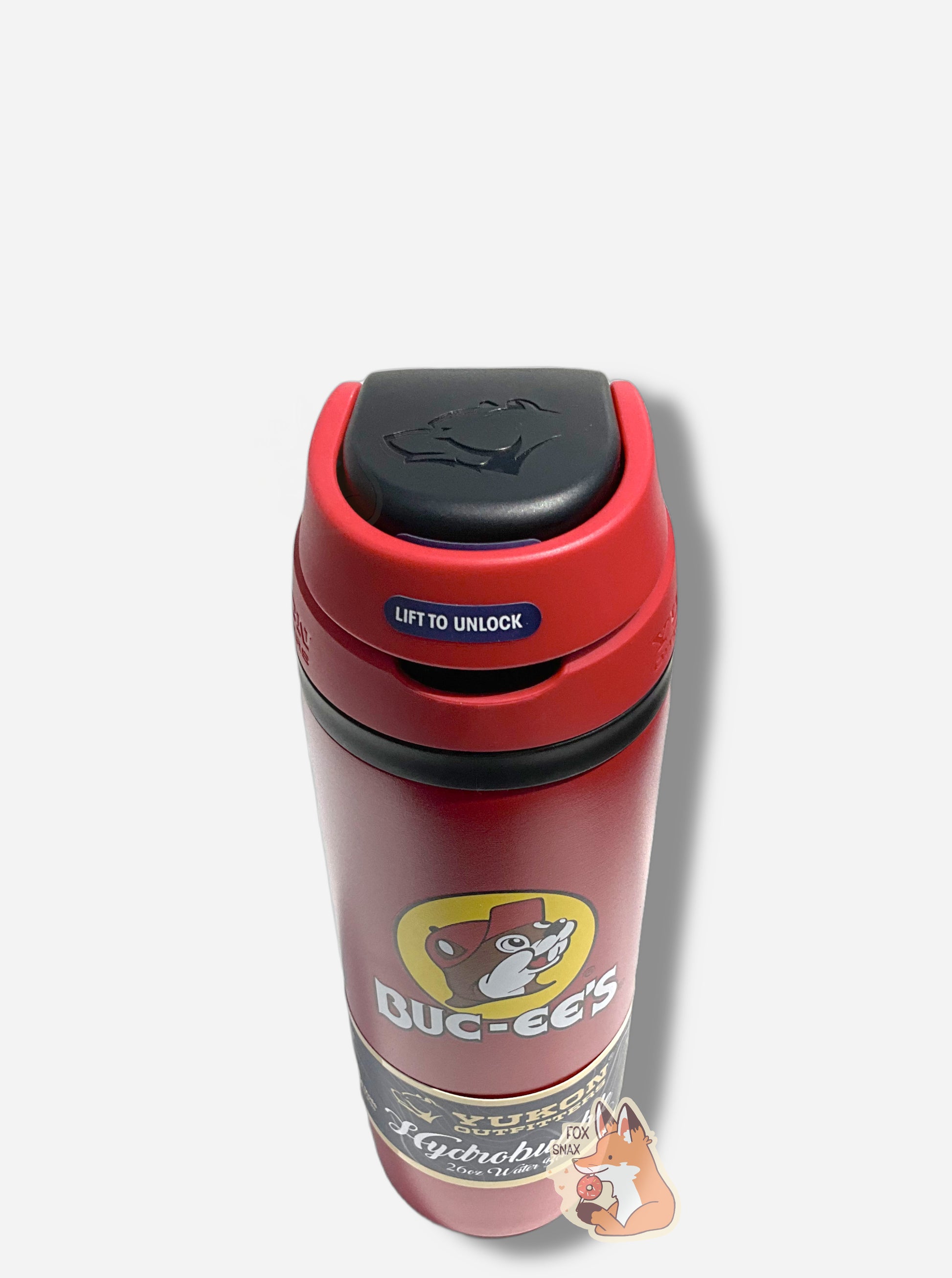 A three-quarters front view of a tall red cylindrical water bottle, with a big picture of Buc-ee The Beaver on it.  It reads BUC-EE'S in a large white circular font.  The surface is smooth and red, as it is red paint covering stainless steel.  The small plastic handle is closed.  The top is black and red plastic, and is closed.  On the top of the handle, the Yukon Outfitters logo - a grumpy bear head - can be seen. A paper wrapper around the bottom reads: Yukon Outfitters.  Hydrobubble 26oz Water Bottle.