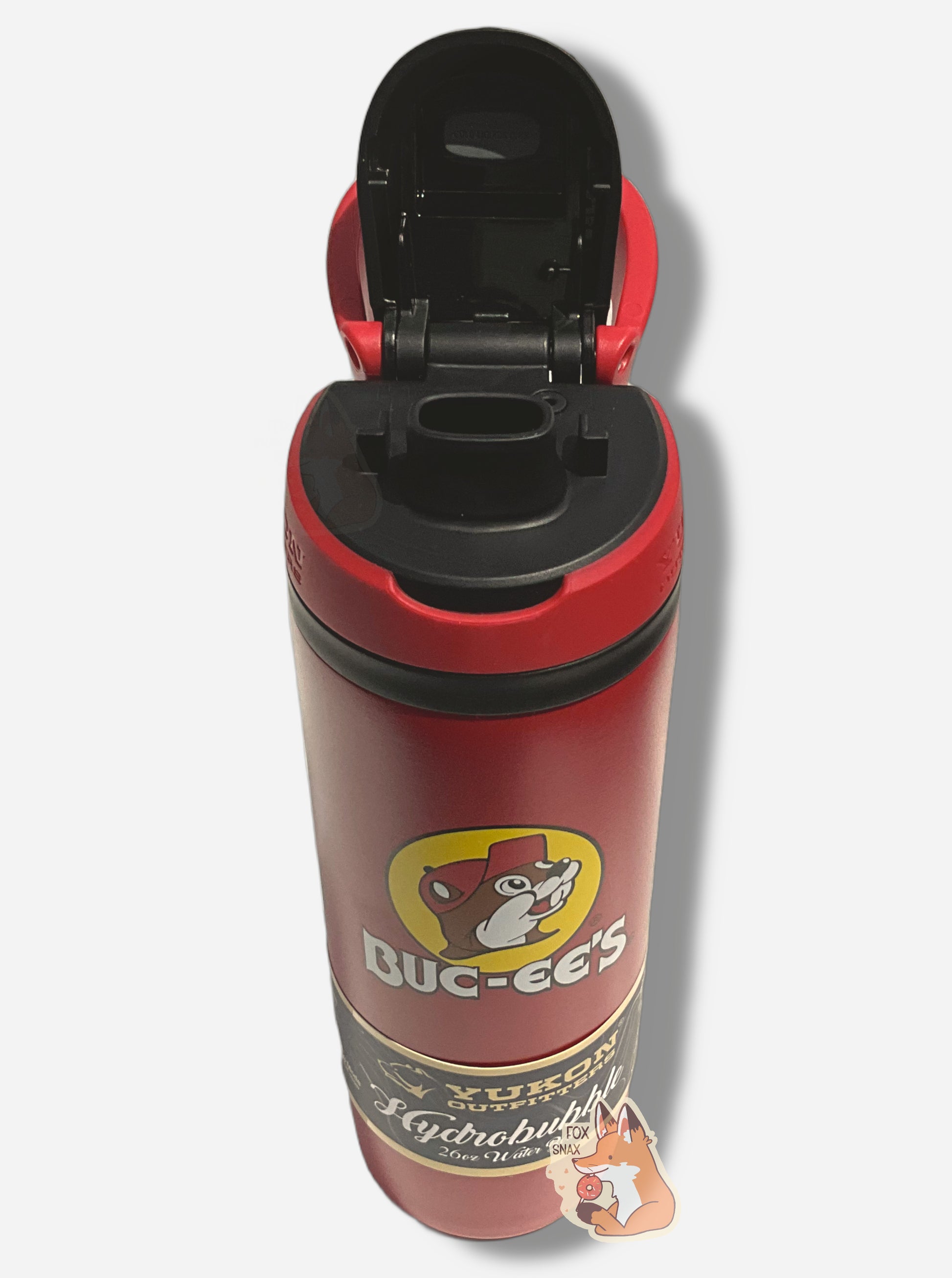 A three-quarters front view of a tall red cylindrical water bottle, with a big picture of Buc-ee The Beaver on it.  It reads BUC-EE'S in a large white circular font.  The surface is smooth and red, as it is red paint covering stainless steel.  The small plastic handle is open, and behind it, the top is open, showing off the oval-shaped Sip & Chug black plastic drinking port.  A paper wrapper around the bottom reads: Yukon Outfitters.  Hydrobubble 26oz Water Bottle.