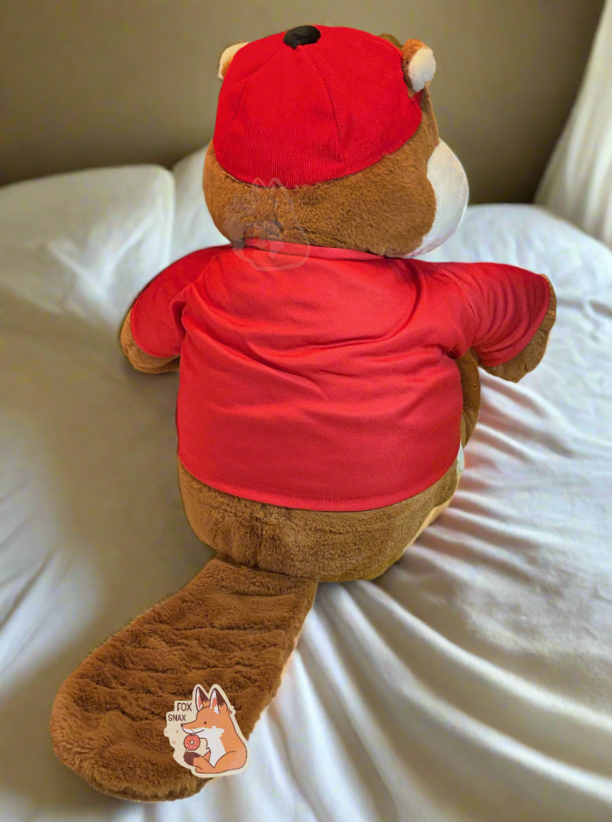 A picture of the back of an adorable Buc-ee the Beaver plush.  The plush is of Buc-ee, wearing a red Buc-ee's shirt and red ball cap, and is looking to the left.  Buc-ee has a large brown fuzzy beaver tail, and is covered in plush brown and white stuffed animal "fur".  The plush can be seen sitting on a bed with warm white covers and yellow/orange lighting.