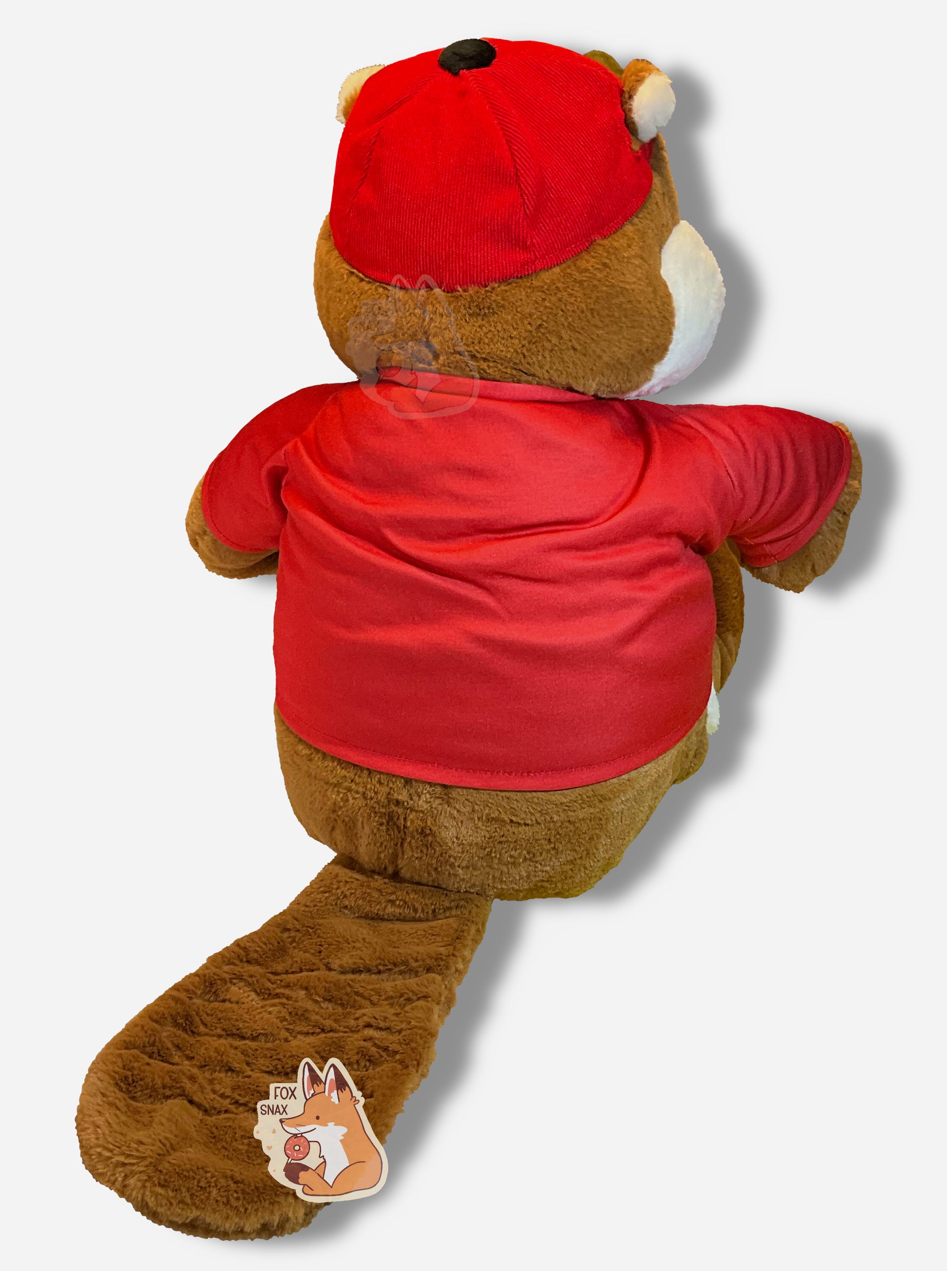 A picture of the back of an adorable Buc-ee the Beaver plush.  The plush is of Buc-ee, wearing a red Buc-ee's shirt and red ball cap, and is looking to the left.  Buc-ee has a large brown fuzzy beaver tail, and is covered in plush brown and white stuffed animal "fur".