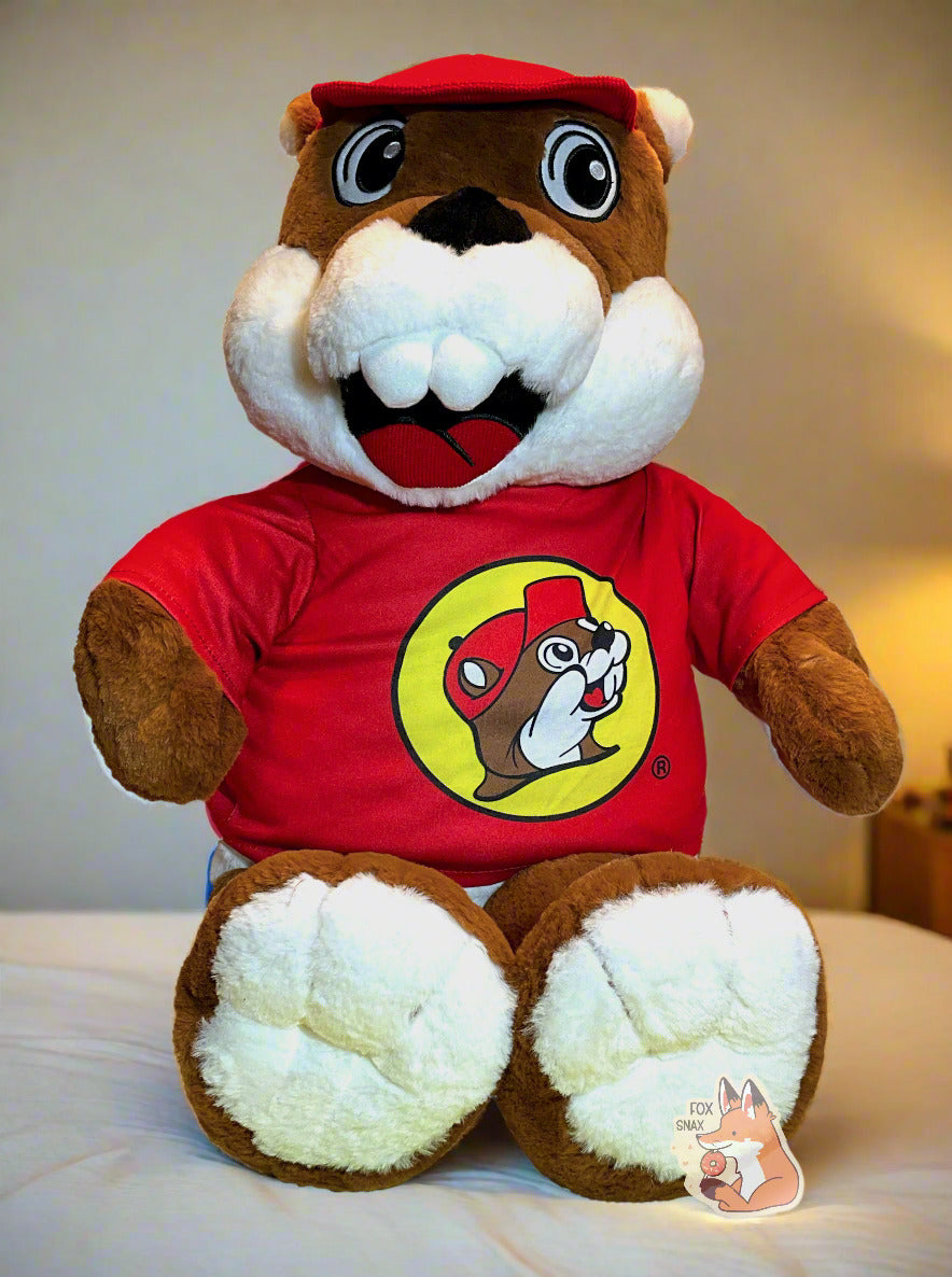 A picture of an adorable Buc-ee the Beaver plush.  The plush is of Buc-ee, wearing a red Buc-ee's shirt and red ball cap, and is looking to the left.  It is roughly two feet tall and covered in plush brown and white stuffed animal "fur".  The plush can be seen sitting on a bed with warm white covers and yellow/orange lighting.
