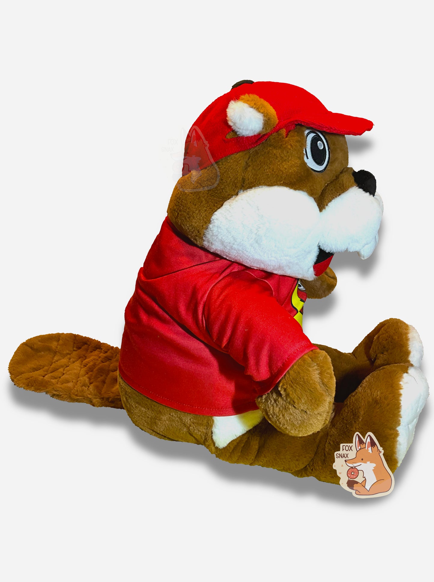 A picture of the side of an adorable Buc-ee the Beaver plush.  The plush is of Buc-ee, wearing a red Buc-ee's shirt and red ball cap, and is looking to the left.  Buc-ee has a large brown fuzzy beaver tail, and is covered in plush brown and white stuffed animal "fur".