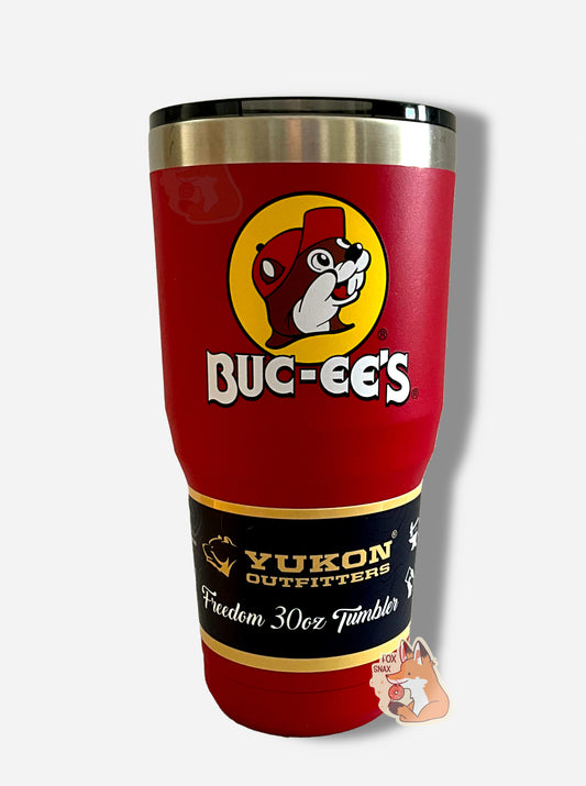 A picture of a tall, red, tumbler, with a big picture of Buc-ee The Beaver on it.  The surface is smooth and red, as it is red paint covering stainless steel.  There is no handle.  The top is plastic.  The top of the mug is lined with a stainless steel band.  A paper wrapper around the bottom reads: Yukon Outfitters.  Freedom 30 oz Tumbler.