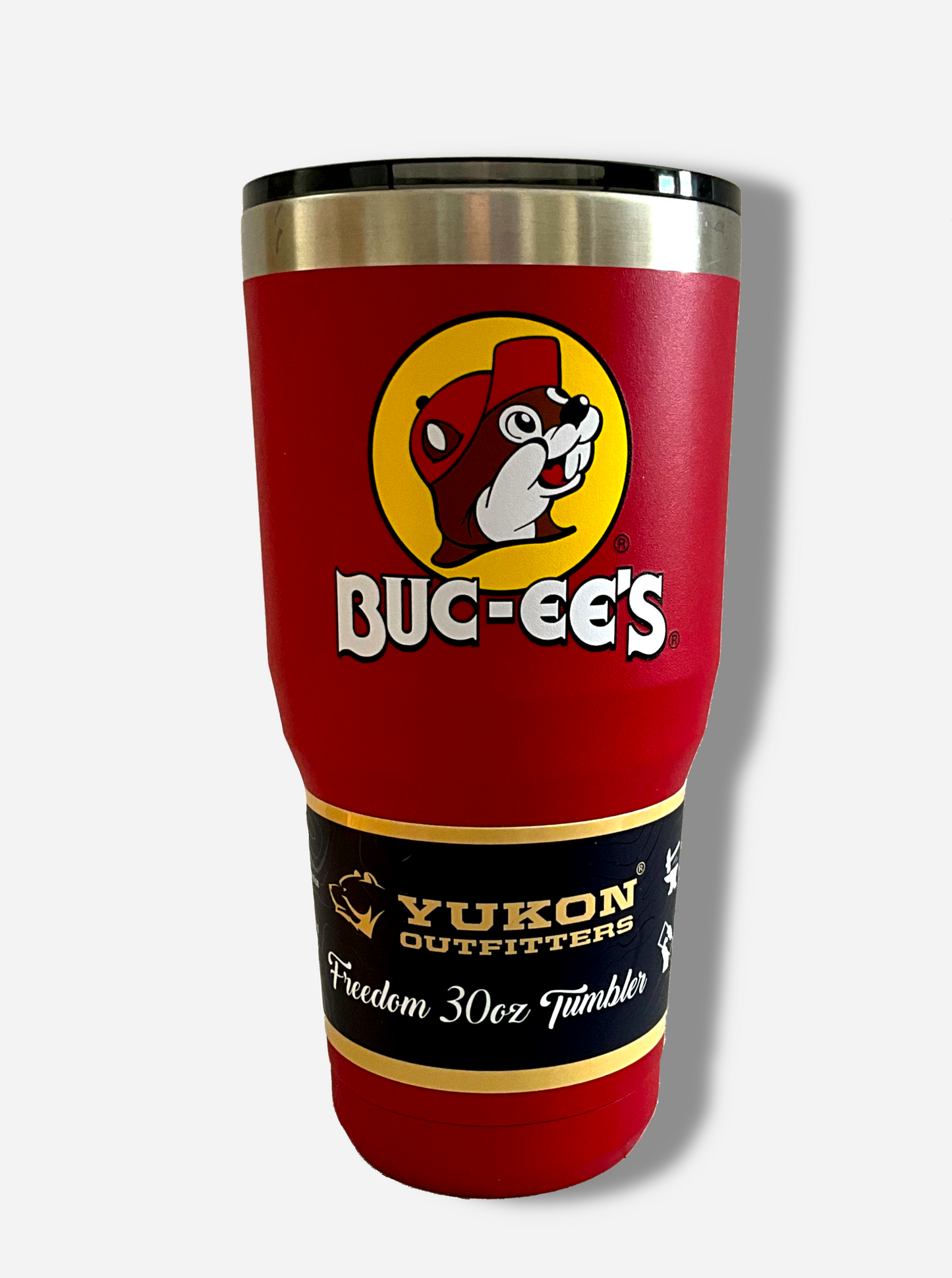 A picture of a tall, red, tumbler, with a big picture of Buc-ee The Beaver on it.  The surface is smooth and red, as it is red paint covering stainless steel.  There is no handle.  The top is plastic.  The top of the mug is lined with a stainless steel band.  A paper wrapper around the bottom reads: Yukon Outfitters.  Freedom 30 oz Tumbler.