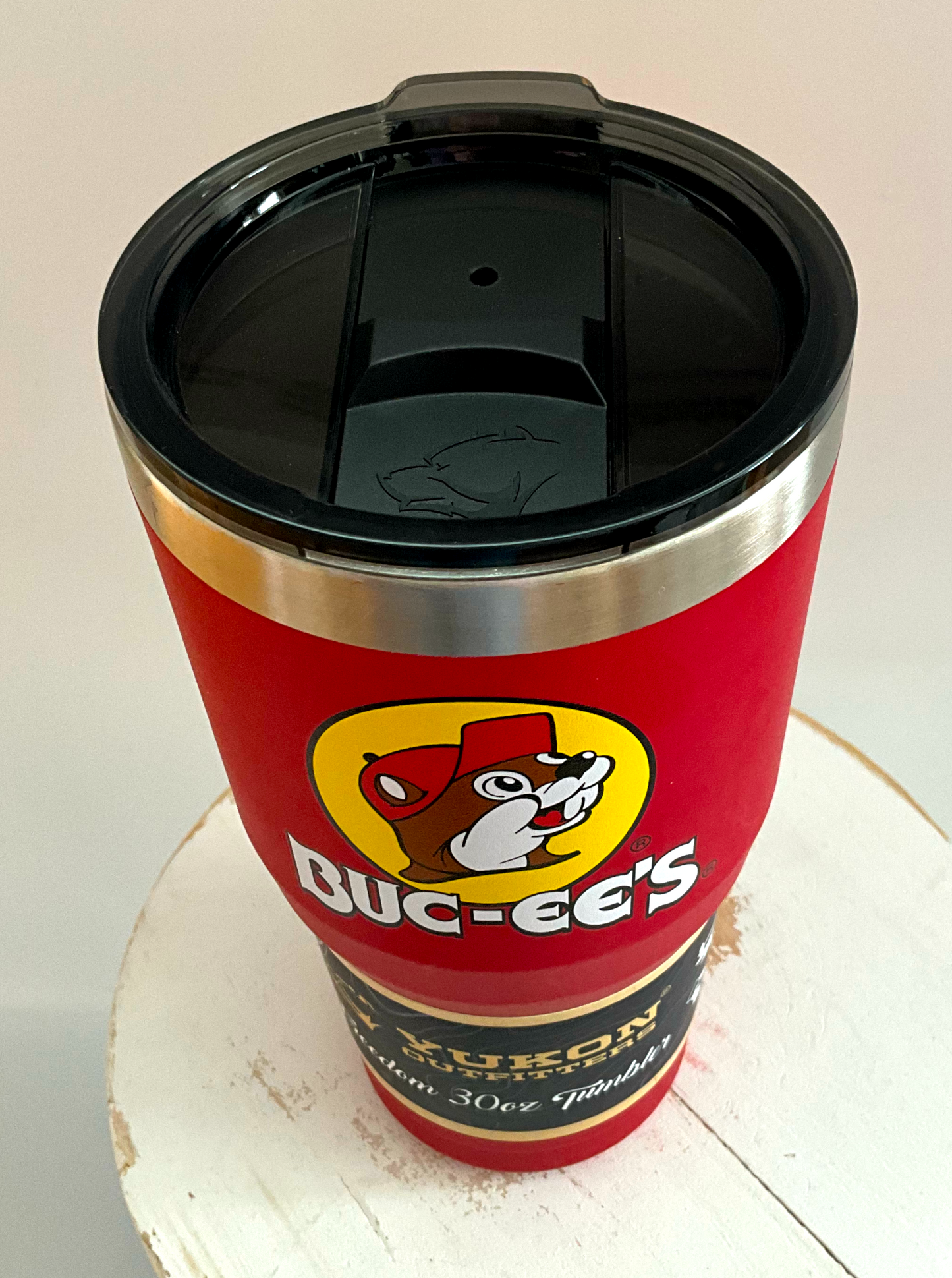 A three-quarters view of a picture of a tall, red, tumbler, with a big picture of Buc-ee The Beaver on it.  The surface is smooth and red, as it is red paint covering stainless steel.  There is no handle.  The top is black plastic, and shows a slider in the middle with the imprint of the Yukon bear's head on it.  The top of the mug is lined with a stainless steel band.  A paper wrapper around the bottom reads: Yukon Outfitters.  Freedom 30 oz Tumbler.  The tumbler is sitting on a white circular stool. 