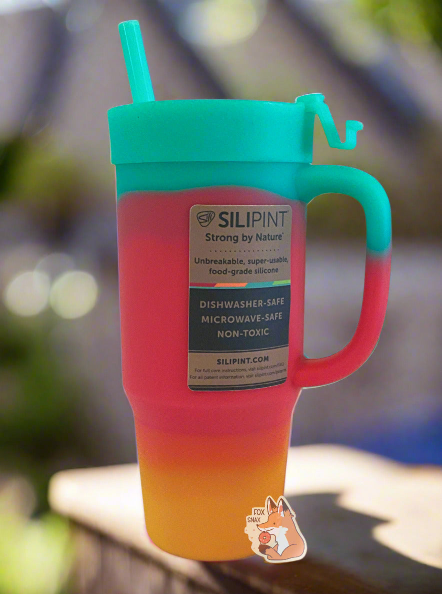 A picture of the back of a tall, rainbow colored tumbler.  The surface starts as orange-yellow on the bottom, fading to red in the middle, then blue/green at the top.  The entire mug is made of silicone, including the top and the straw.  A brown sticker, in black font on the back, reads "SILIPINT, Strong By Nature, Unbreakable, super-usable, food-grade silicone.  Dishwasher-safe, Microwave-safe, Non-toxic.  The tumbler is sitting on an edge of a hot tub; the tub's blue water can be seen in the background.