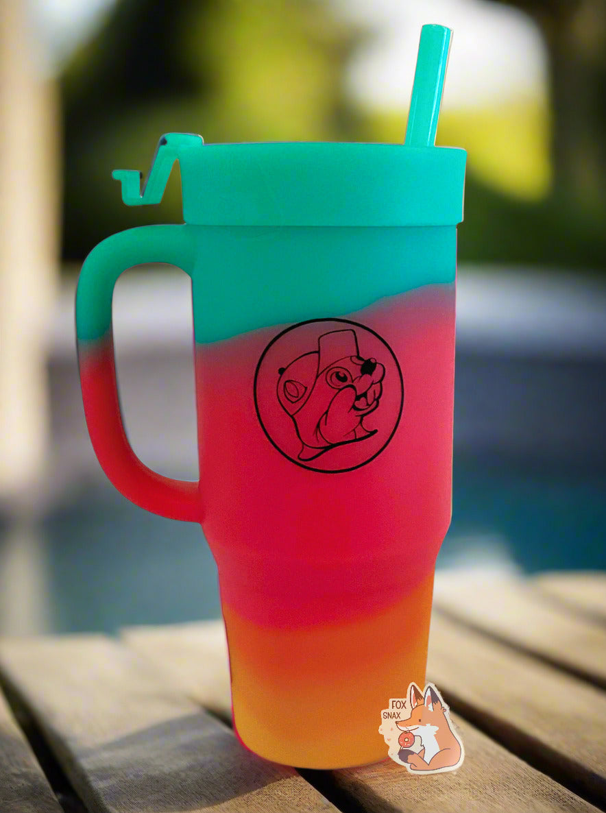 A picture of a tall, rainbow colored tumbler with a big outline of Buc-ee The Beaver on it in black.  The surface starts as orange-yellow on the bottom, fading to red in the middle, then blue/green at the top.  The entire mug is made of silicone, including the top and the straw.  The tumbler is sitting on an edge of a hot tub; the tub's blue water can be seen in the background.