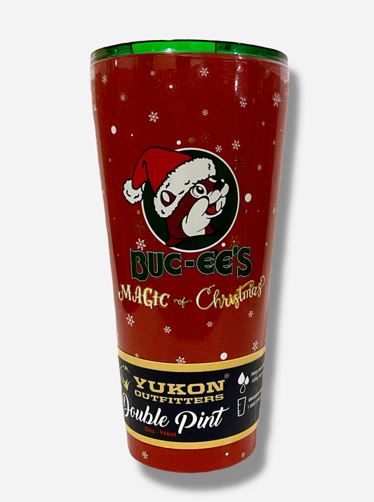 A picture of a red double pint tumbler, with a dark red background full of white snowflakes.  The middle of the tumbler has the Buc-ee's logo, but with Buc-ee The Beaver wearing a Santa hat.  Beneath it reads BUC-EE'S in dark green text, then underneath that, MAGIC OF CHRISTMAS in fancy gold/yellow font.  The top of the tumbler is green clear plastic, with a red slide for different beverage dispenser amounts.  The label on the bottom reads YUKON OUTFITTERS, then DOUBLE PINT 32 OZ