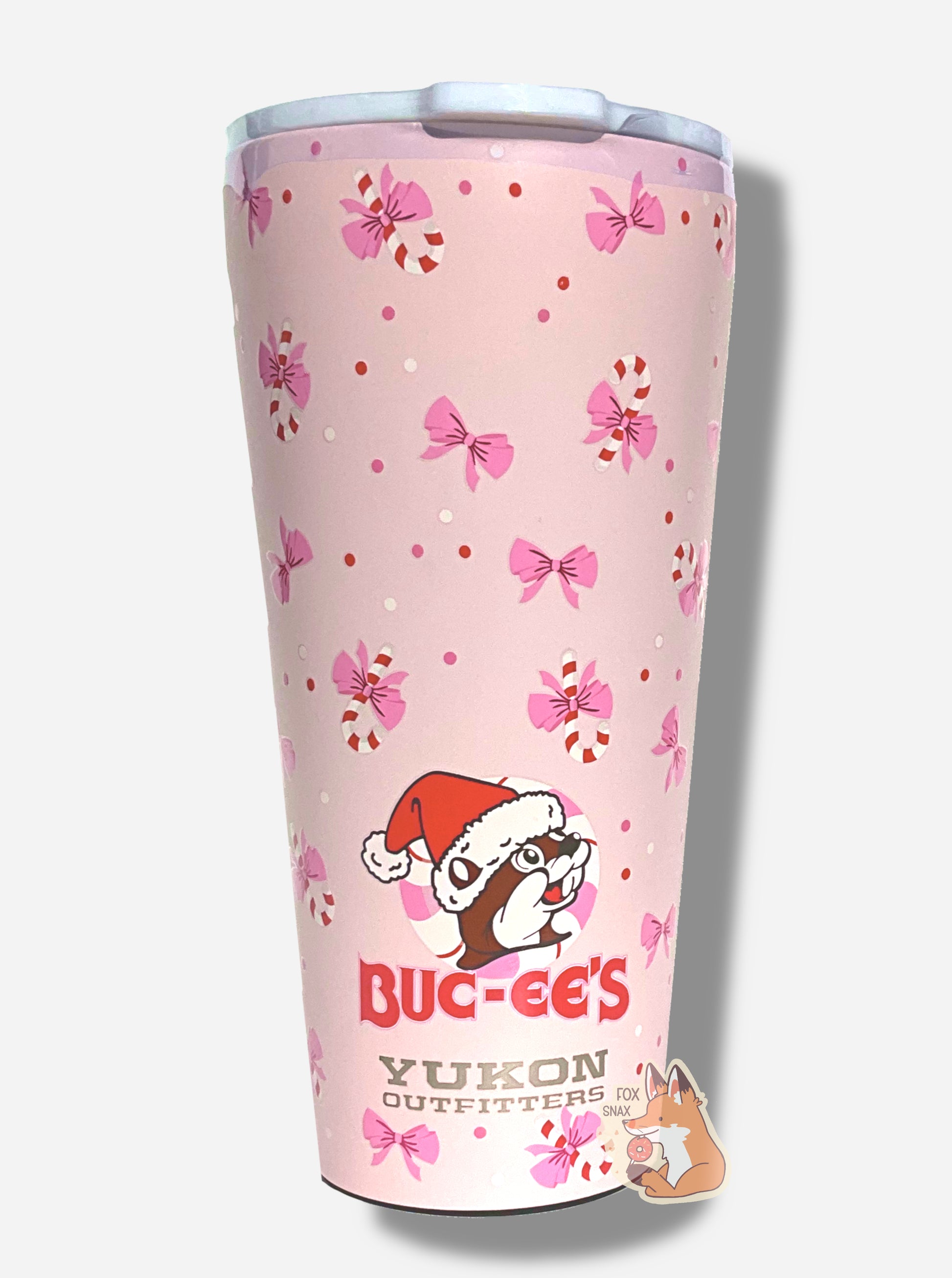 A picture of a pink double pint tumbler, with a light pink background full of bows and candy canes.  The middle bottom of the tumbler has the Buc-ee's logo, but with Buc-ee The Beaver wearing a Santa hat, framed against a peppermint.  The label on the bottom reads BUC-EE'S in red text, then underneath that, YUKON OUTFITTERS.