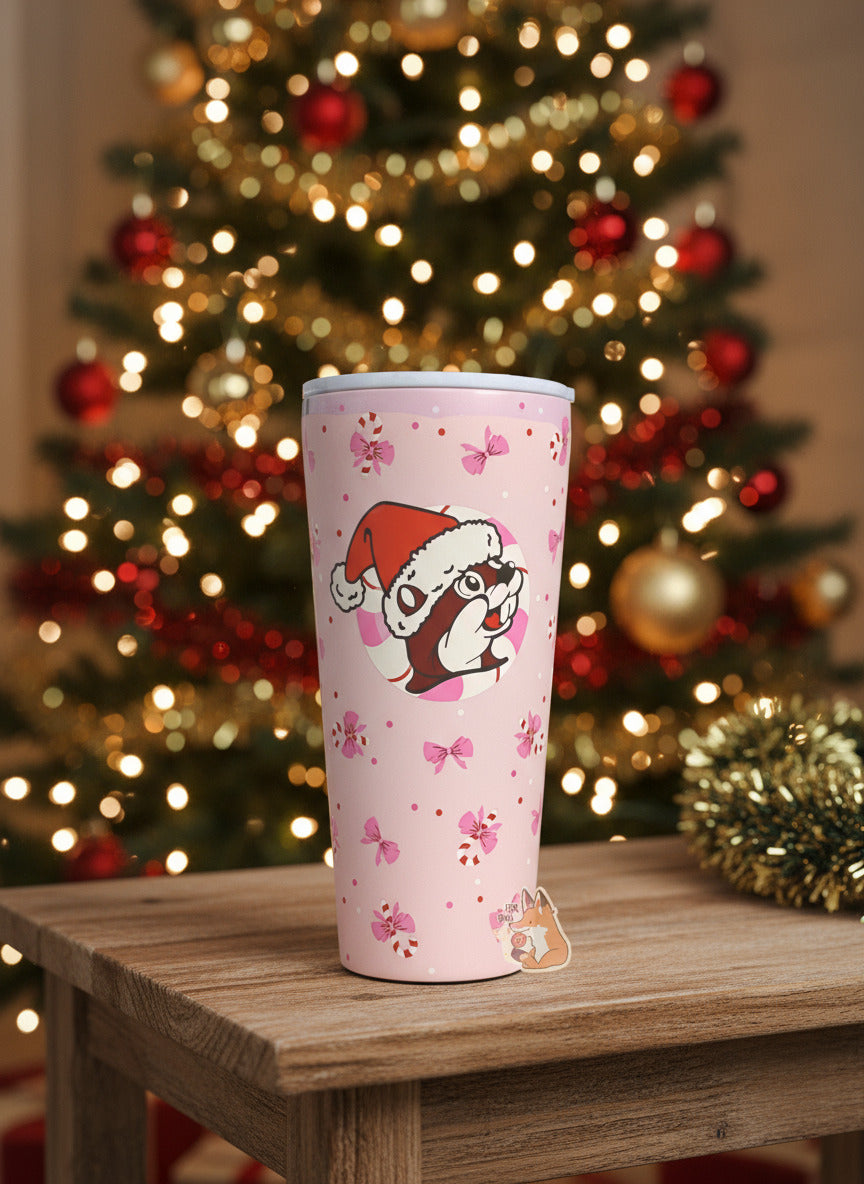 A picture of a pink double pint tumbler, with a light pink background full of bows and candy canes.  The middle of the tumbler has the Buc-ee's logo, but with Buc-ee The Beaver wearing a Santa hat, framed against a red and white peppermint.  The top of the tumbler is white, with a pink slide for different beverage dispenser amounts.  The tumbler is sitting on a wooden end table, against a beautiful Christmas tree background.