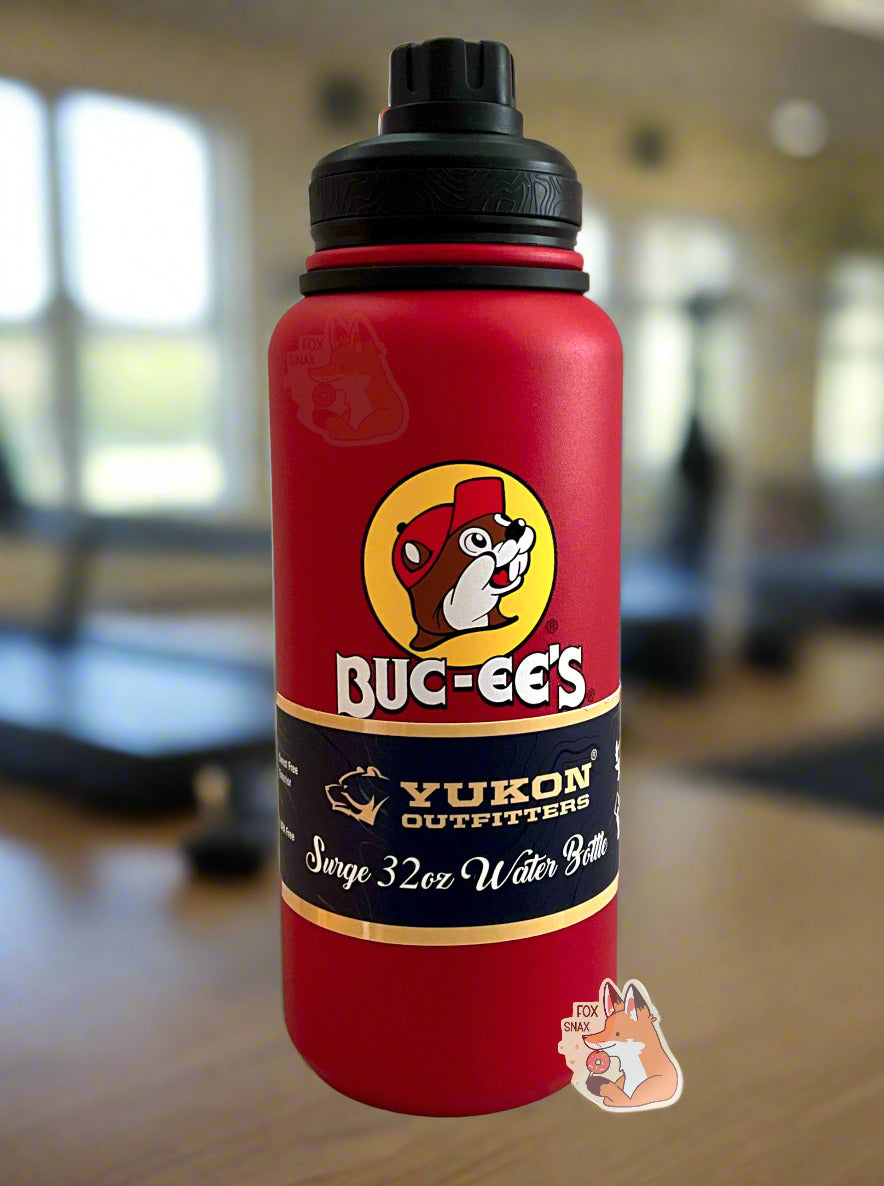 A picture of a tall red cylindrical water bottle, with a big picture of Buc-ee The Beaver on it.  The surface is smooth and red, as it is red paint covering stainless steel.  There is no handle.  The top is a screw on plastic knob with a keychain handle.  A paper wrapper around the bottom reads: Yukon Outfitters.  Surge 32 oz Water Bottle.  The background is a wood-floored, clean, well polished gymnasium; treadmills can be seen in the background.