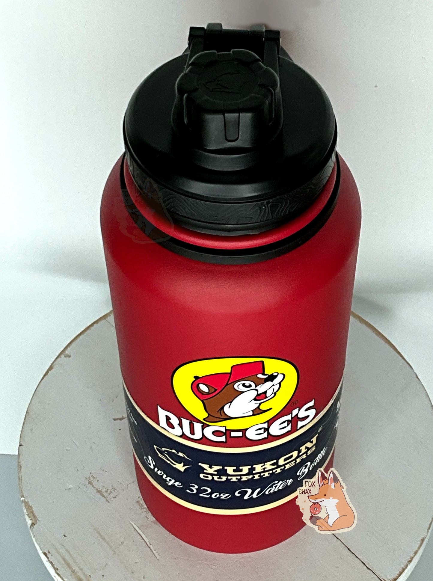 A three quarters view of a tall red cylindrical water bottle, with a big picture of Buc-ee The Beaver on it.  The surface is smooth and red, as it is red paint covering stainless steel.  There is no handle.  The top is a screw on plastic knob with a keychain handle.  A paper wrapper around the bottom reads: Yukon Outfitters.  Surge 32 oz Water Bottle.  The water bottle it sitting on a white painted stool.