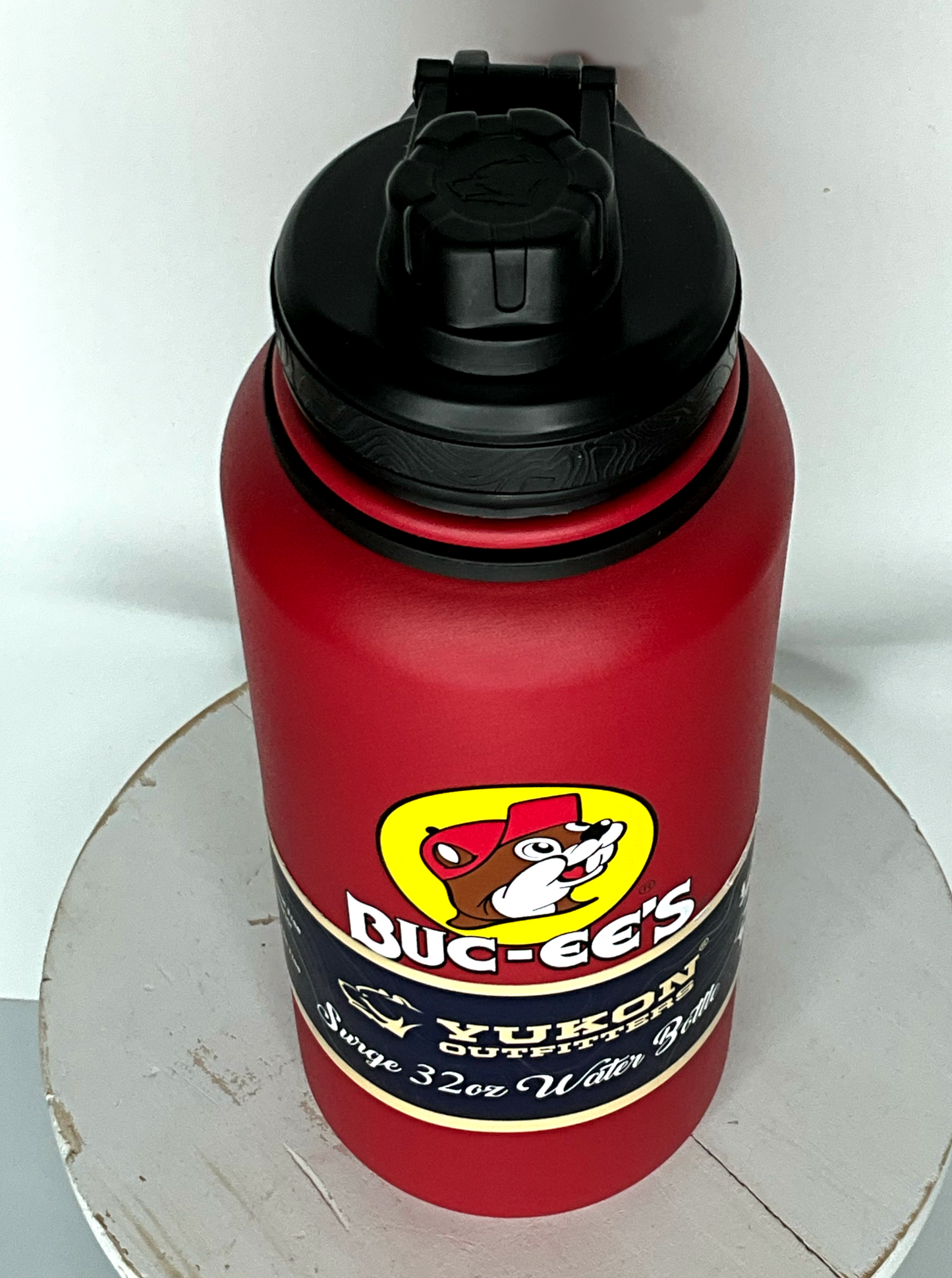 A three quarters view of a tall red cylindrical water bottle, with a big picture of Buc-ee The Beaver on it.  The surface is smooth and red, as it is red paint covering stainless steel.  There is no handle.  The top is a screw on plastic knob with a keychain handle.  A paper wrapper around the bottom reads: Yukon Outfitters.  Surge 32 oz Water Bottle.  The water bottle it sitting on a white painted stool.