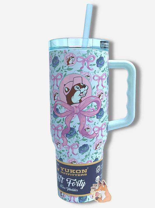 A picture of a tall, light blue, sorta skinny tumbler, with a big picture of Buc-ee The Beaver on it.  Buc-ee's hat, and the circle behind him, is in light pink, and is framed by a large light pink bow.  The surface is dotted with Buc-ee's logos, the pink bows, and dark blue flowers.  It is matte paint in covering stainless steel.  The handle, top, and straw are light blue (the top and straw are transparent) and plastic.  A paper wrapper around the bottom reads: Yukon Outfitters.  Fit Forty 40 oz Tumbler.