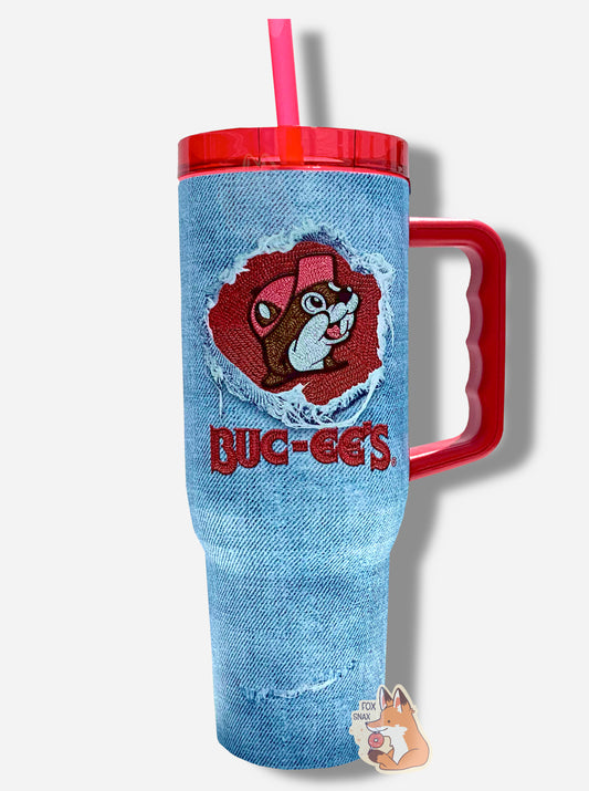 A picture of a tall, faded denim blue, sorta skinny tumbler, with a big picture of Buc-ee The Beaver on it in the middle, inside of a circular denim "rip".  The surface is bumpy and light blue, as if made out of denim (it's not; it's metal).  Several "rips" mar the surface; they show the white stitching underneath.  The tumbler  is light blue paint covering stainless steel.  The handle, top, and straw are red plastic. 