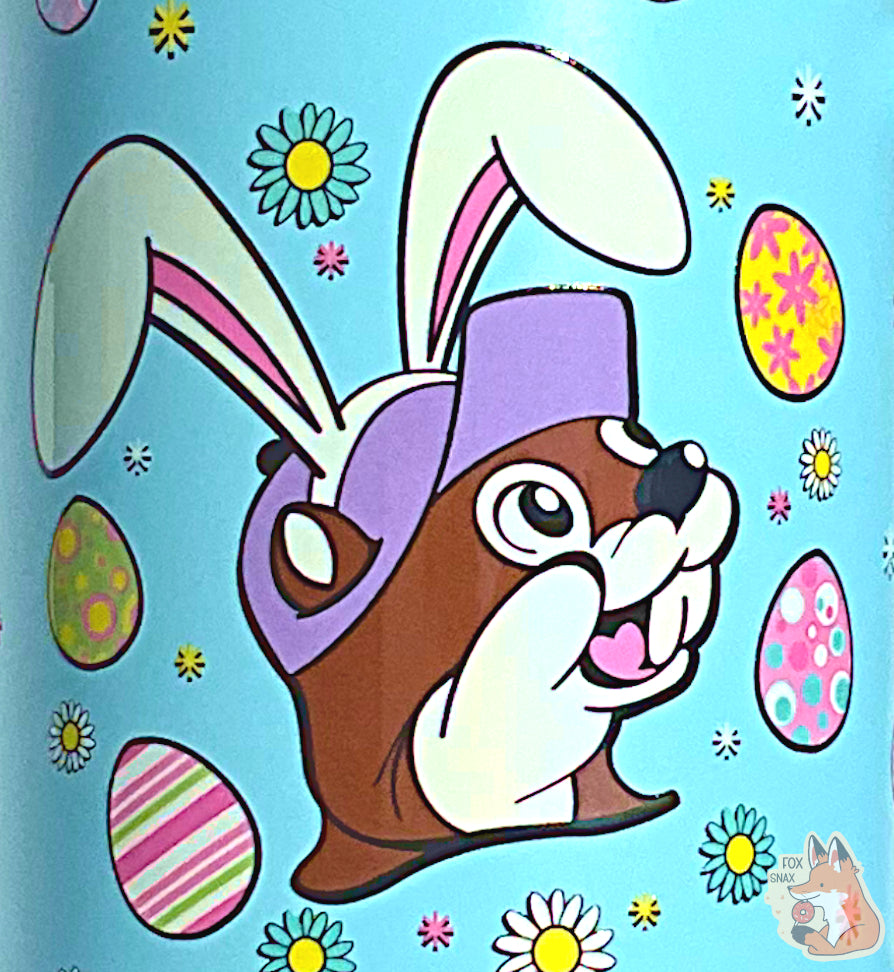 A closeup of a picture of a tall, light blue, sorta skinny tumbler, with a picture of Buc-ee the Beaver's head, wearing white Easter Bunny ears.  Above and below his head,  are a series of Easter eggs dyed in fantastic patterns and colors, as well as daisy-like flower heads in different petal colors, with a yellow center.  The surface is light blue, as it is paint covering stainless steel.  