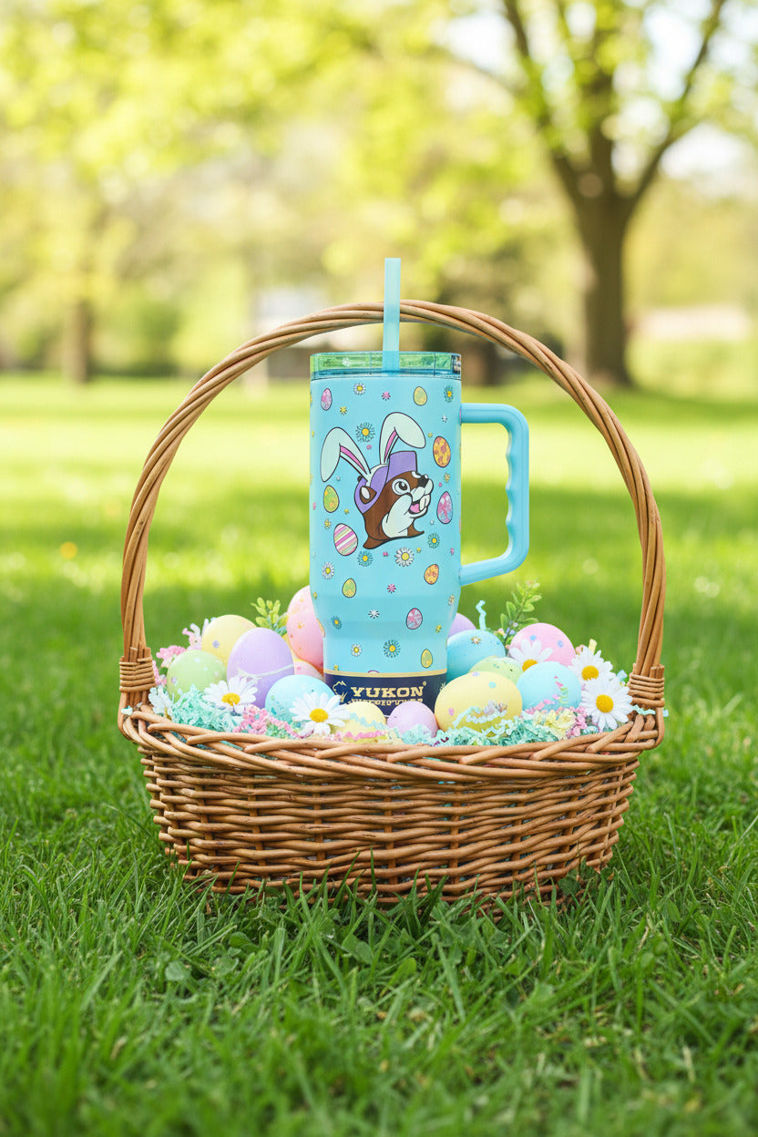 A picture of a tall, light blue, sorta skinny tumbler, with a picture of Buc-ee the Beaver's head, wearing white Easter Bunny ears.  Above and below his head,  are a series of Easter eggs dyed in fantastic patterns and colors, as well as daisy-like flower heads in different petal colors, with a yellow center.  The tumbler is sitting in the middle of an wooden Easter basket; the basket is in a park in a field of spring grass.