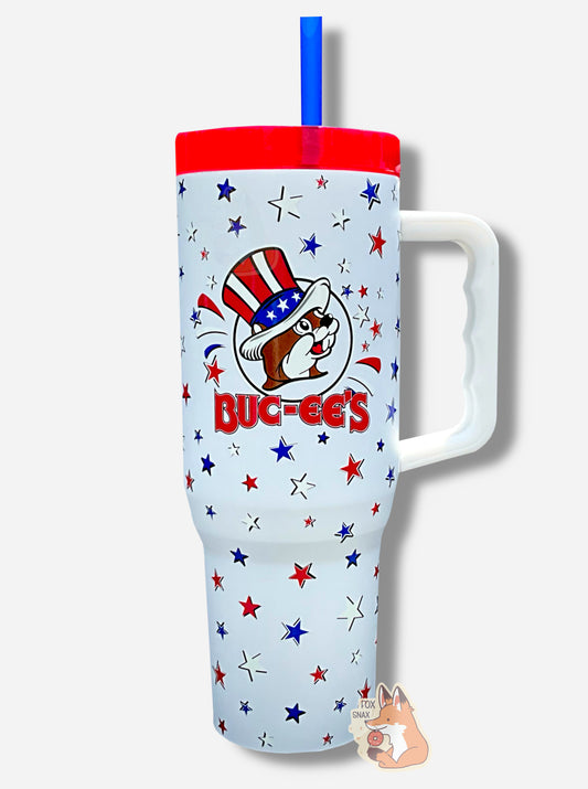 A picture of a tall, white, sorta skinny tumbler, with a big picture of Buc-ee The Beaver on it wearing a red, white, and blue Uncle Sam hat and the words BUC-EES in red.  The surface is smooth and white, covered with red, white, and blue stars.  It is white  paint covering stainless steel.  The handle (white), top (red) and straw (blue) are plastic. This is a  Yukon Outfitters Fit Forty 40 oz Tumbler.  