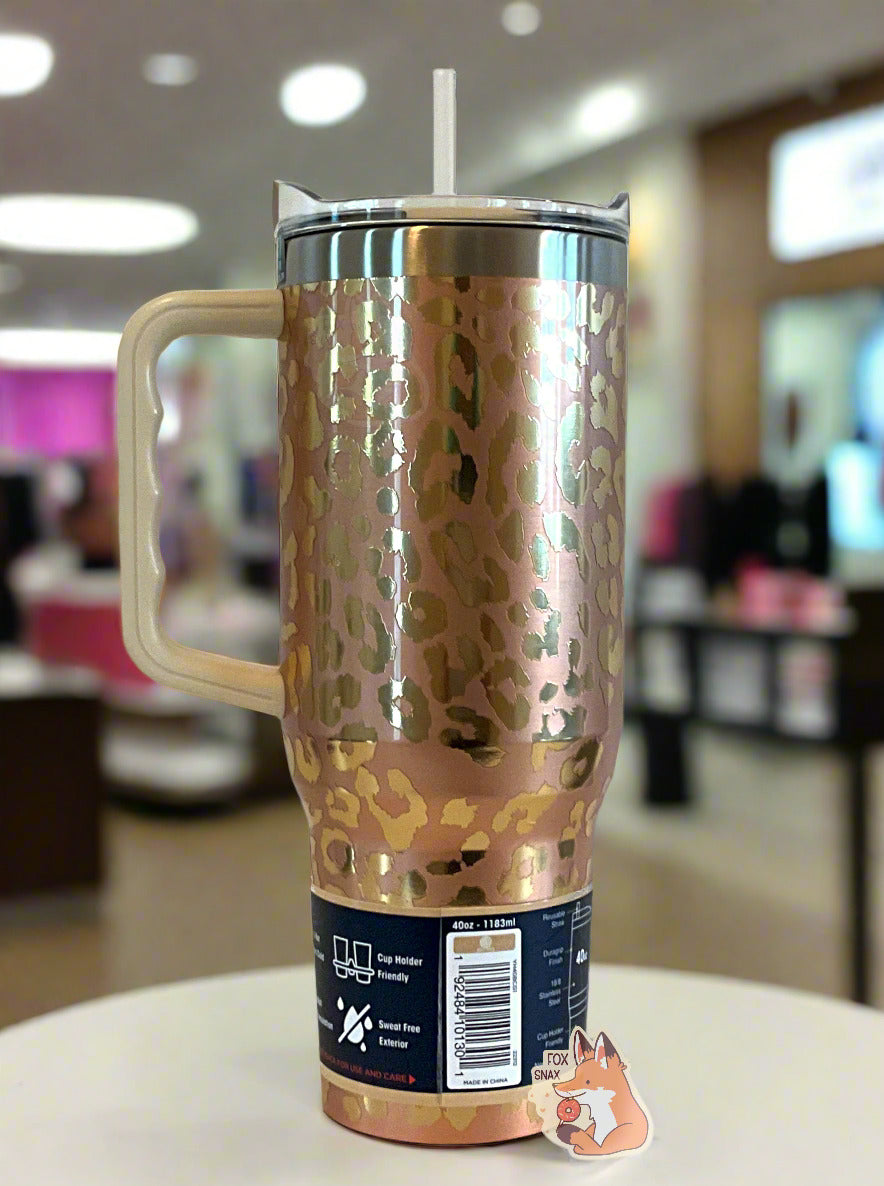 A picture of the back of a tall, gold, sorta skinny tumbler.  The surface is a leopard print, as it is gold paint covering stainless steel.  The handle, top, and straw are plastic.  The top of the mug is lined with a stainless steel band.  A paper wrapper around the bottom has a diagram that reads 40 oz, next to the barcode.  The background shows the tumbler sitting on a white table in a well-lit, upscale mall.