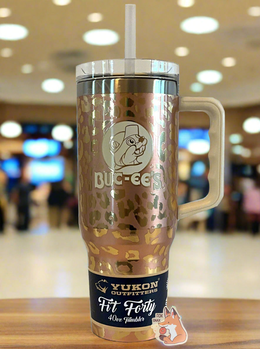 A picture of a tall, gold, sorta skinny tumbler, with a big picture of Buc-ee The Beaver on it.  The surface is a leopard print, as it is gold paint covering stainless steel.  The handle, top, and straw are plastic.  The top of the mug is lined with a stainless steel band.  A paper wrapper around the bottom reads: Yukon Outfitters.  Fit Forty 40 oz Tumbler.  The background shows the tumbler sitting on a bench in a well-lit, upscale mall.