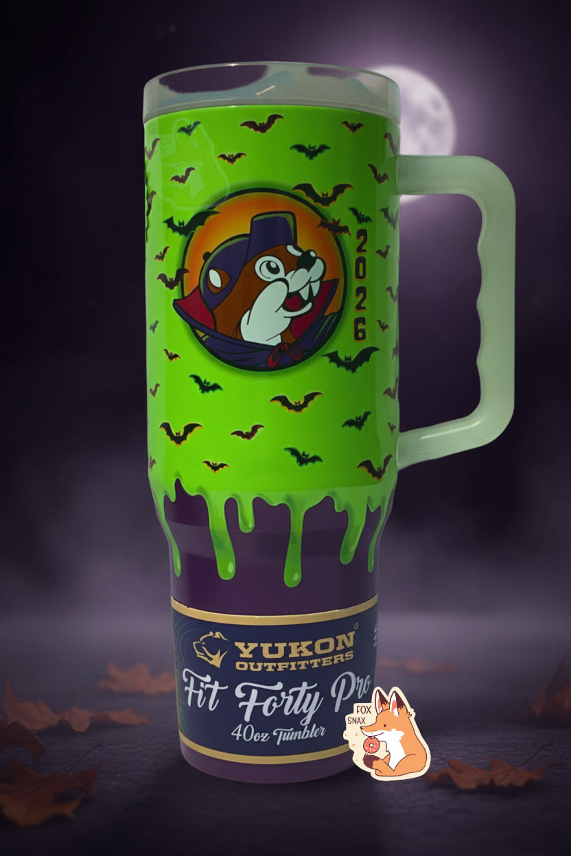 A picture of a tall, green and purple sorta skinny tumbler, with a giant picture of Buc-ee in the middle, wearing a red/blue vampire cowl. The background behind him appears to be a orange/yellow full moon, and he has, interestingly, two fangs instead of the usual beaver overbite. The background is green slime 'dripping' down the tumbler, with black bats against that background.  The background is of a spooky, dark leaf-covered surface with fog rising from it; a full moon can be seen in the distance.