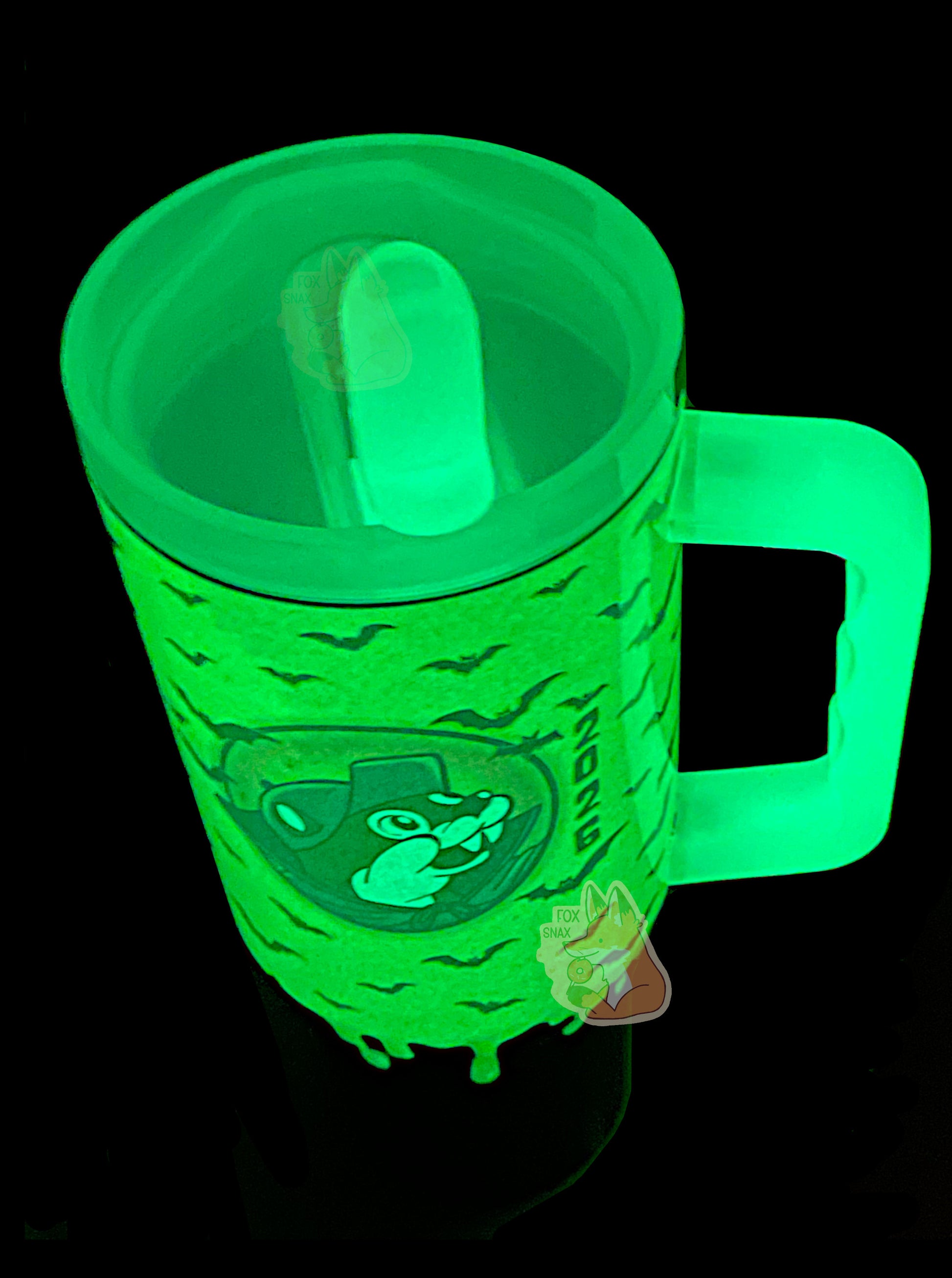 A three-quarters top down view of a tall, dark sorta skinny tumbler, with a giant picture of Buc-ee in the middle, complete with vampire cowl and fangs. All of this is glowing bright green, as it is glow-in-the-dark paint. Silhouettes of bats against the green slime area can be seen; the formerly purple area on the bottom is now black and doesn't glow. The handle and top are glow in the dark plastic.