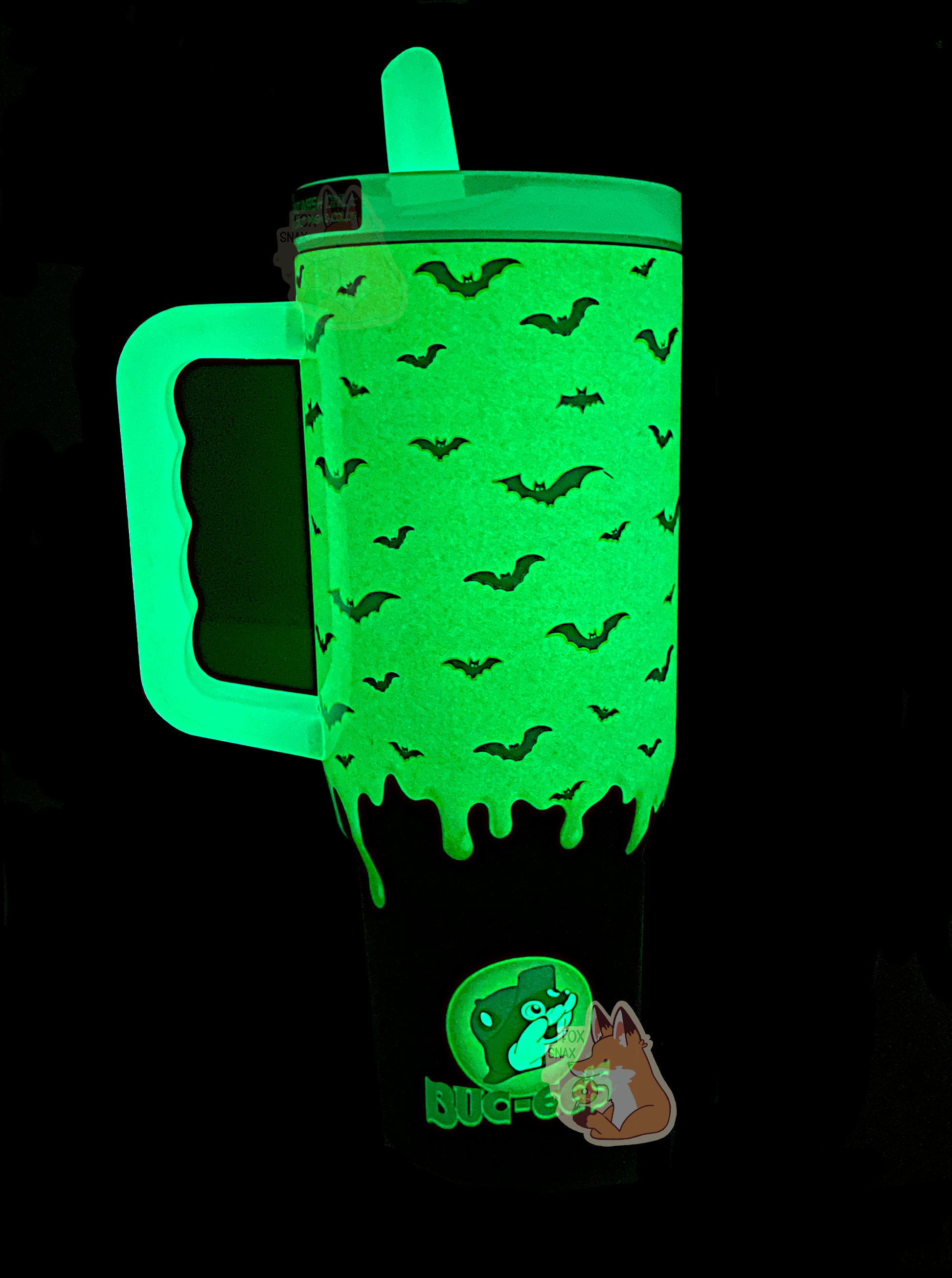 A picture of the back of a tall, green and purple sorta skinny tumbler. All of this is glowing bright green, as it is glow-in-the-dark paint. Silhouettes of bats against the green slime area can be seen; the formerly purple area on the bottom is now black and doesn't glow. On the bottom is a regular (non-vampiric) Buc-ee's logo, glowing green.  The handle and top are glow in the dark plastic.