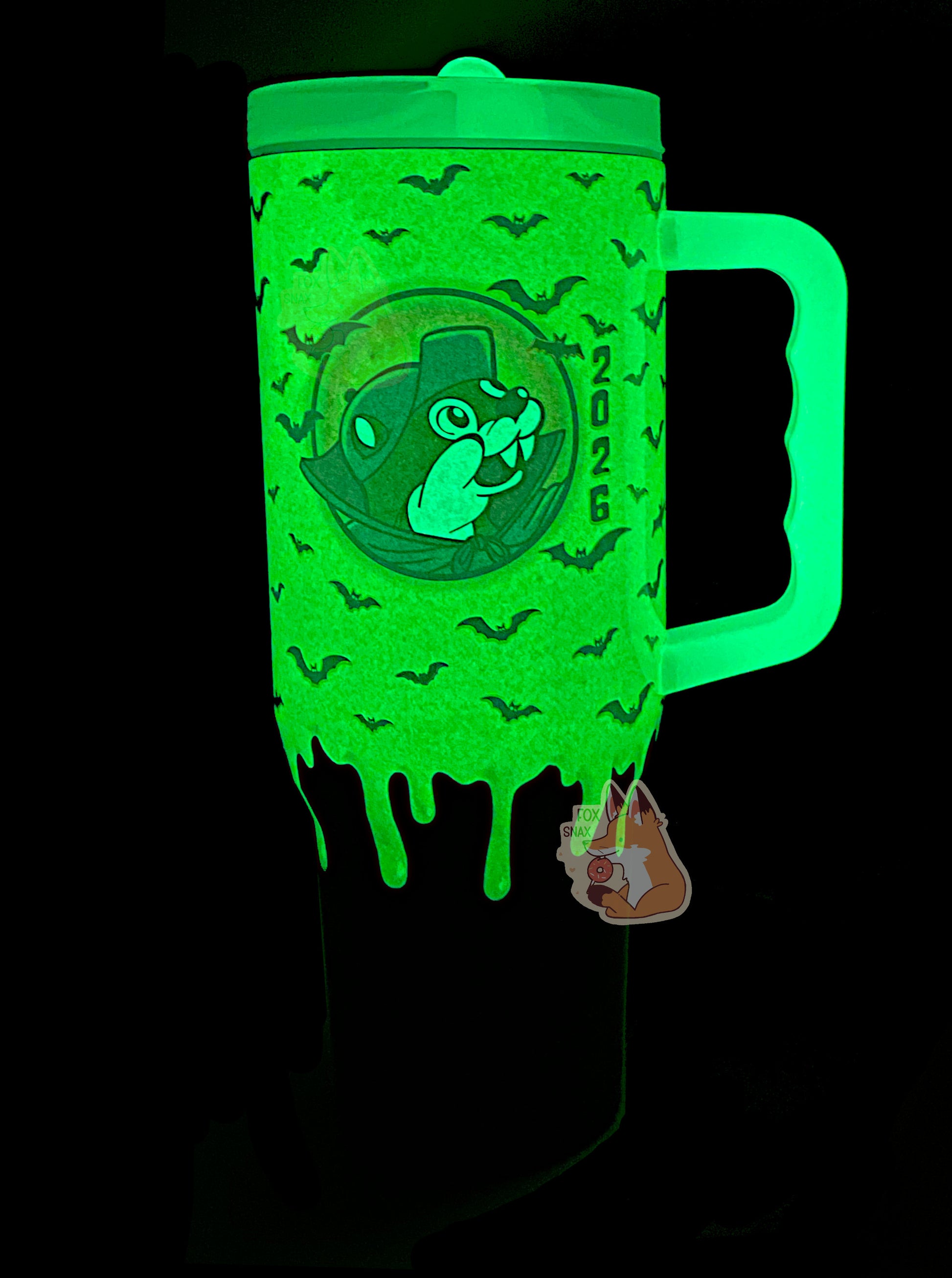 A picture of a tall, green and purple sorta skinny tumbler, with a giant picture of Buc-ee in the middle, complete with vampire cowl and fangs. All of this is glowing bright green, as it is glow-in-the-dark paint. Silhouettes of bats against the green slime area can be seen; the formerly purple area on the bottom is now black and doesn't glow. The handle and top are glow in the dark plastic.