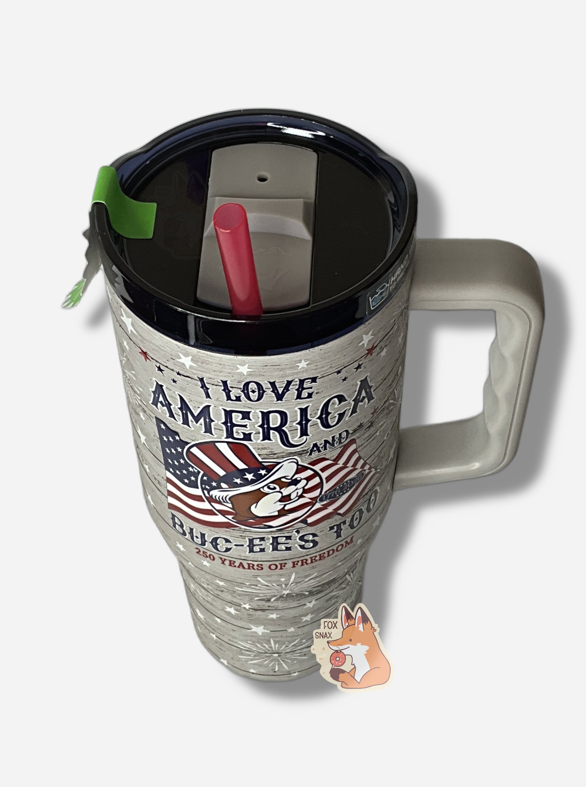 A three-quarters view of the front of a tall, grey "woodgrain", sorta skinny tumbler, with a big picture of Buc-ee The Beaver on it wearing a red, white, and blue Uncle Sam hat and the words I LOVE AMERICA AND BUC-EE'S TOO in red.  The surface is slightly textured, grey,  covered with white stars and white exploding fireworks.  Underneath the main text, it reads 250 YEARS OF FREEDOM.  It is grey paint covering stainless steel. 