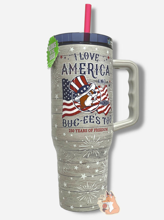 A picture of a tall, grey "woodgrain", sorta skinny tumbler, with a big picture of Buc-ee The Beaver on it wearing a red, white, and blue Uncle Sam hat and the words I LOVE AMERICA AND BUC-EE'S TOO in red.  The surface is slightly textured, grey,  covered with white stars and white exploding fireworks.  Underneath the main text, it reads 250 YEARS OF FREEDOM.  It is grey paint covering stainless steel. The handle (grey), top (blue) and straw (red) are plastic; a Yukon Outfitters Fit Forty 40 oz Tumbler.  