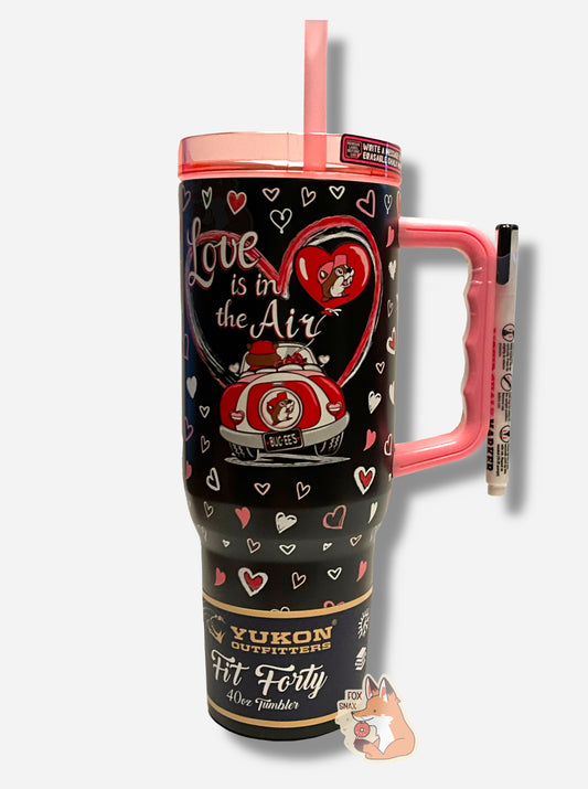 A picture of a tall, skinny, black, 40 oz tumbler.  The lid, handle, and straw are pink (the top is transparent pink).  The body of the tumbler is black, and features the text LOVE IS IN THE AIR in a white cursive font, with a pink/white heart surrounding it.  Beneath it is a picture of Buc-ee The Beaver, from the back  driving a red and white convertible with a bouquet of roses.  The rest of the tumbler is decorated with white and pink hearts.  A white chalk marker is taped to the handle for use on back.